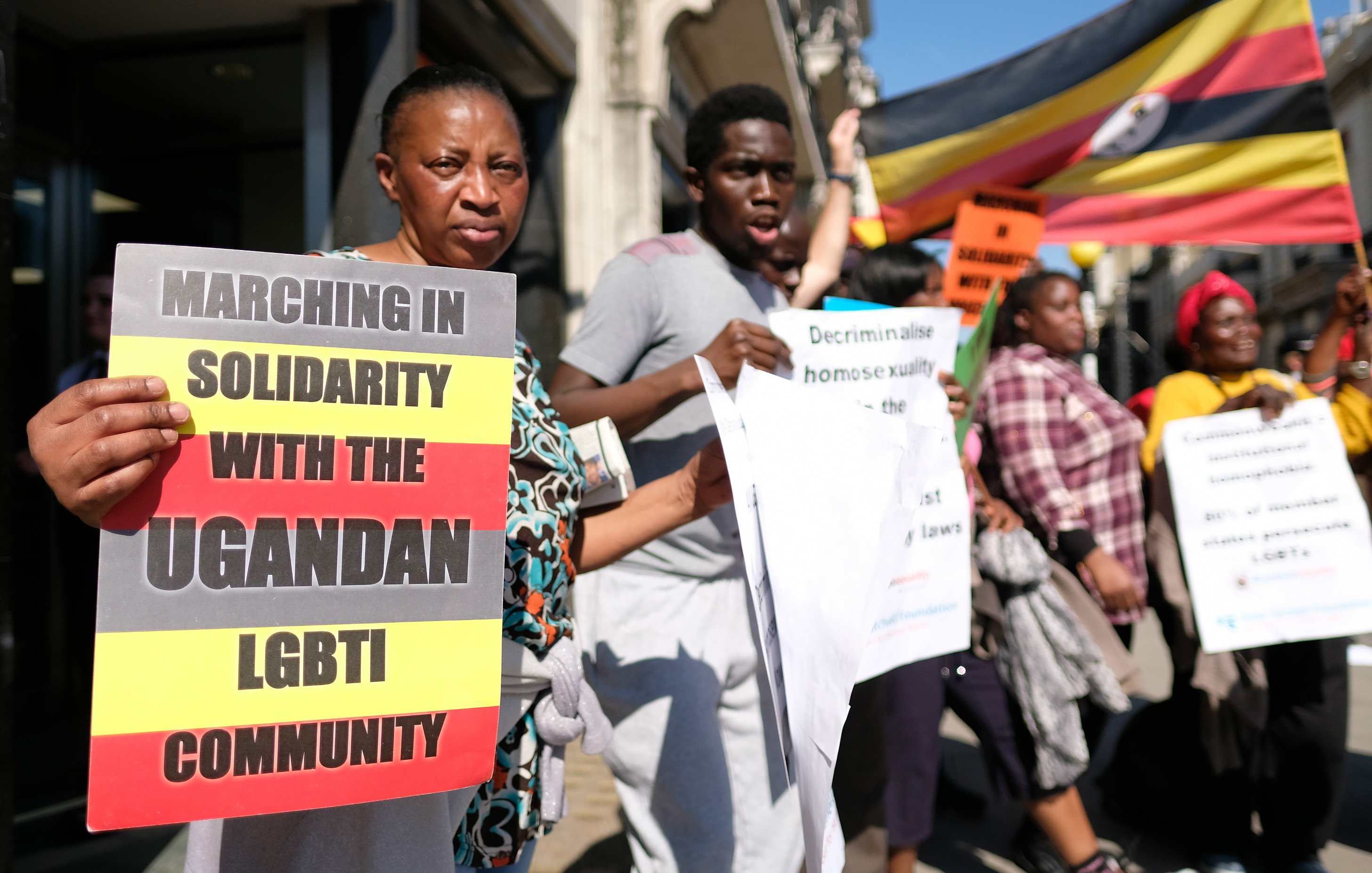 Dans cette image, un groupe de personnes participe à une marche de solidarité en faveur de la communauté LGBTI ougandaise. Les manifestants tiennent des pancartes avec des messages tels que "Marchons en solidarité avec la communauté LGBTI ougandaise" et "Décriminalisez l'homosexualité". L'ambiance est énergique et déterminée, et le groupe affiche une grande diversité. En arrière-plan, on peut voir un drapeau de l'Ouganda flapper, créant un contraste visuel avec les affiches et les vêtements des participants. Les personnes expriment un fort engagement pour les droits humains et l'égalité.