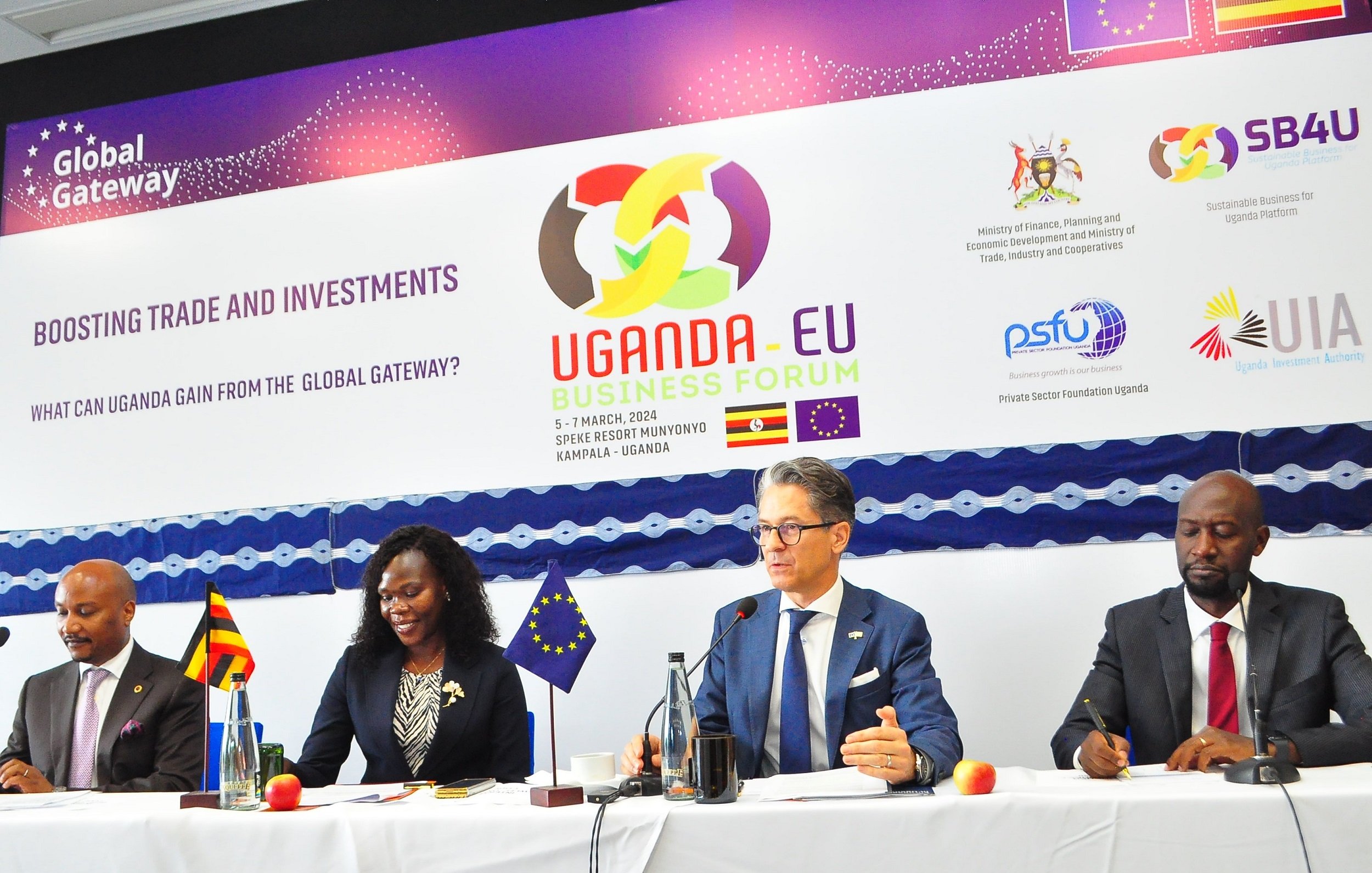L'image montre une scène d'un forum d'affaires intitulé "Uganda-EU Business Forum". Au premier plan, quatre personnes sont assises à une table, chacune ayant un micro devant elle. À gauche, un homme souriant semble représenter l'Ouganda. Ensuite, une femme, également souriante, prend la parole. Au centre, un homme, avec des lunettes et un air sérieux, est en train de s'exprimer. Enfin, à droite, un autre homme, avec une posture attentive, écoute. En arrière-plan, une banderole avec des logos et le titre de l'événement est accrochée. L'ambiance est professionnelle, et l'événement semble axé sur le renforcement des échanges et des investissements entre l'Uganda et l'Union européenne.