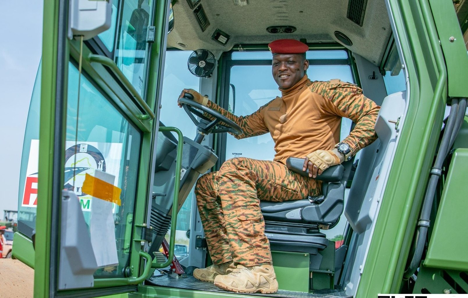 Dans cette image, nous voyons un homme assis à l'intérieur d'un tracteur. Il porte un uniforme couleur sable orné de motifs camouflés et un béret rouge. Son expression est confiante et il sourit, montrant un sentiment de fierté. À l'arrière-plan, on aperçoit d'autres personnes habillées de manière similaire, travaillant dans un environnement agricole. Le ciel est clair et lumineux, suggérant une journée ensoleillée. L'ensemble de la scène dégage une atmosphère de travail collectif et de détermination.