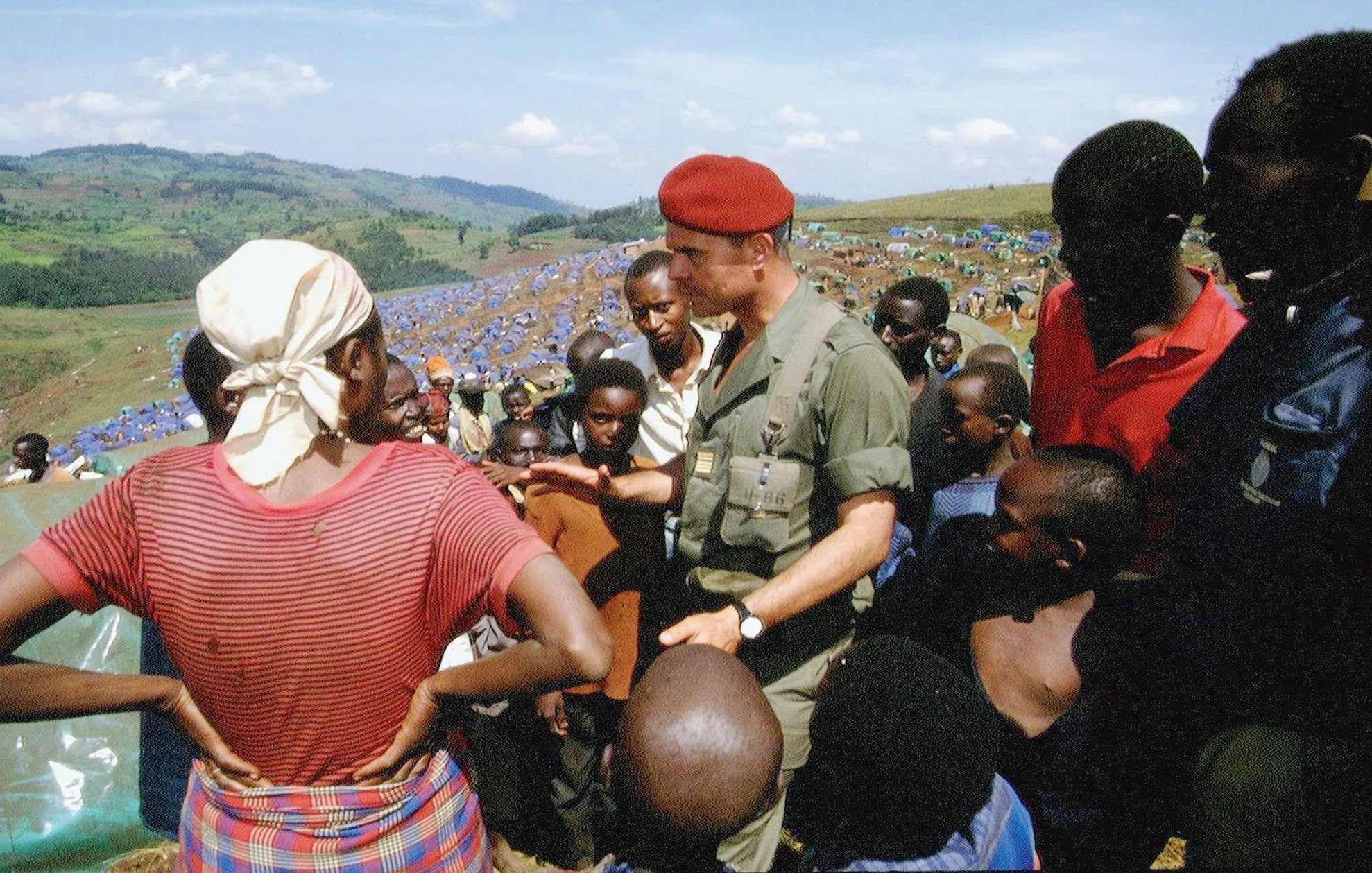 L'image montre un groupe de personnes dans un paysage ouvert et verdoyant. Au centre, un homme en uniforme militaire, portant un béret rouge, discute avec une femme qui porte un foulard et une blouse, le regard tourné vers lui. Elle semble attentive aux explications qu'il donne. Autour d'eux, plusieurs enfants et adultes, principalement d'origine africaine, écoutent avec intérêt. Les visages sont expressifs, certains avec des regards curieux. À l'arrière-plan, on aperçoit une vaste étendue de tentes bleues, indiquant probablement un camp de réfugiés ou de soutien. Le ciel est clair, avec quelques nuages, et les collines verdoyantes offrent une vue panoramique sur la scène. L'atmosphère dégage un sentiment de communauté et d'engagement.