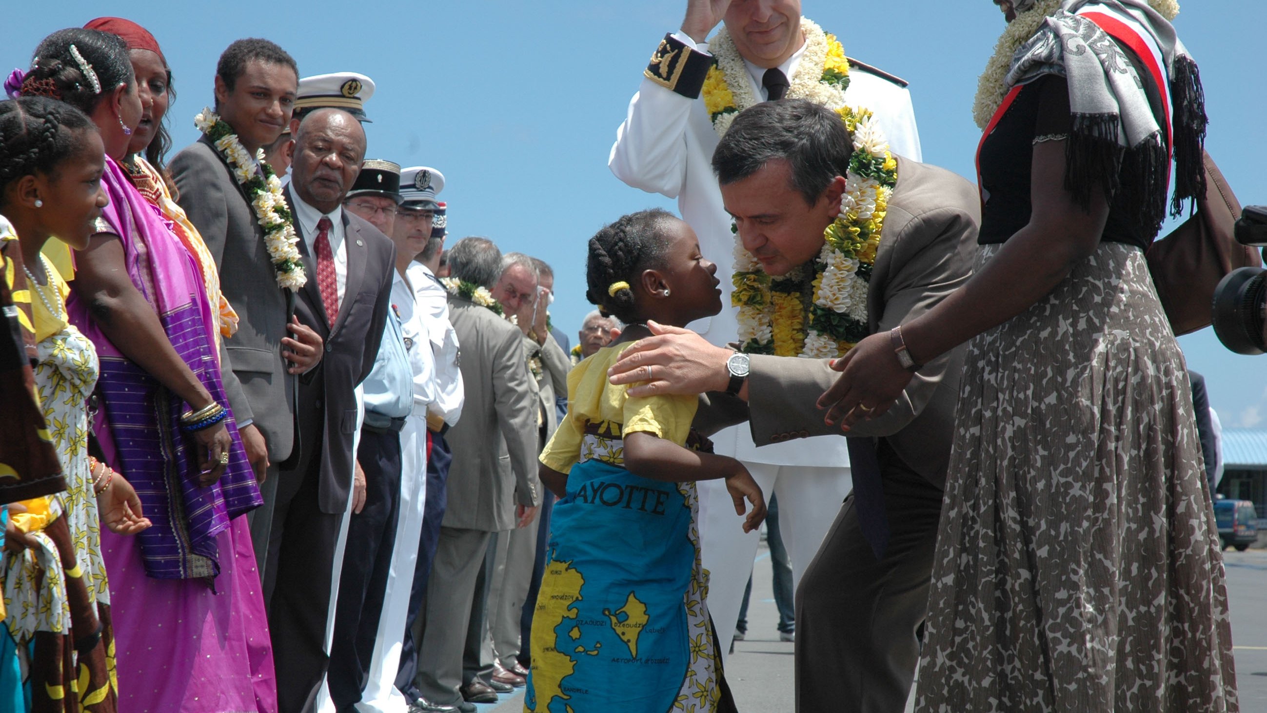 L'image représente une scène d'accueil vibrant sur un aéroport. Au premier plan, un homme en costard se penche pour embrasser une petite fille qui porte une robe colorée et un paréo. La fillette a un grand sourire et semble très joyeuse. Autour d'eux, plusieurs autres personnes, dont des adultes en vêtements traditionnels et militaires, les observent avec des expressions bienveillantes. Certains présents portent des guirlandes de fleurs autour du cou, ajoutant une touche festive à l'atmosphère. En arrière-plan, le ciel est clair, suggérant une journée ensoleillée. L'ensemble de la scène dégage une impression de chaleur et de convivialité.