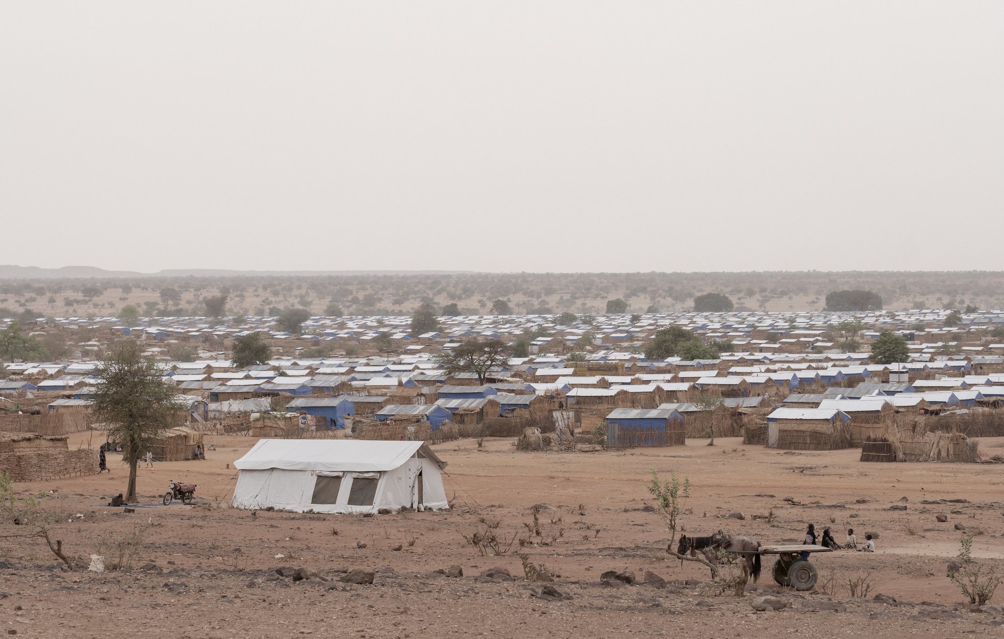 L'image montre un camp de réfugiés. Au premier plan, un enfant assis sur un amas de coussins et de matelas, portant un t-shirt rouge. Il semble pensif, regardant au loin. À côté de lui, un autre enfant est assis sur une petite table, vêtu d'un t-shirt bleu. L'environnement est poussiéreux, avec plusieurs tentes blanches en arrière-plan. On peut également apercevoir des objets éparpillés, comme des cartons et des bidons, évoquant une vie temporaire et difficile. L'atmosphère semble mélancolique et montre une réalité de vulnérabilité.