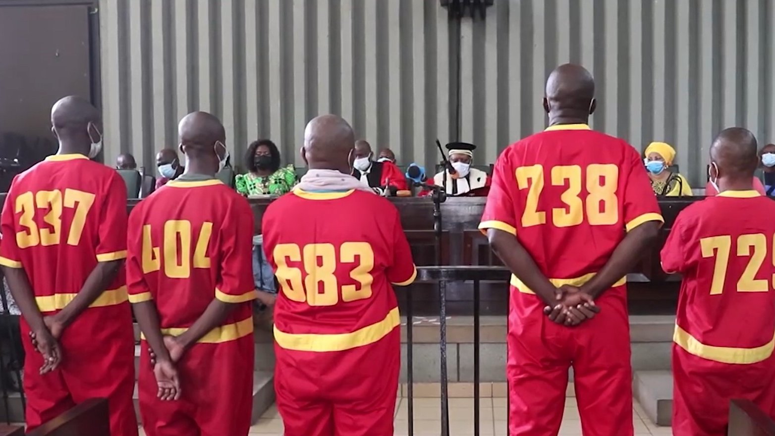 L'image montre un groupe de personnes dans un tribunal. Quatre hommes, portant des uniformes rouges numérotés au dos (337, 404, 683, 238, et 72), se tiennent debout, de dos, face à un juge et à un panel de personnes assises en face d'eux. L'atmosphère est sérieuse, et tous les participants portent des masques. Les murs du tribunal sont rayés, et on peut voir des membres de l'auditoire, y compris des femmes en vêtements colorés. L'ambiance générale évoque une situation judiciaire formelle.