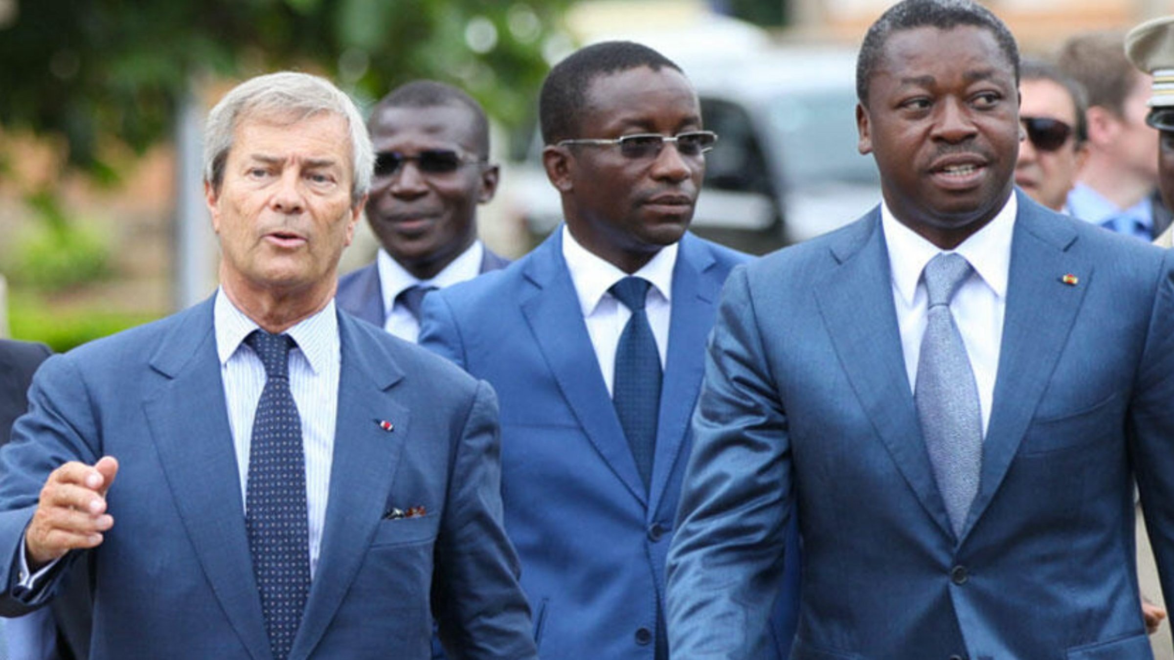 L'image montre un groupe d'hommes marchant ensemble. Le premier homme, à gauche, porte un costume bleu clair et une cravate à rayures. Il semble engagé dans la conversation. Le deuxième homme, au centre, porte aussi un costume bleu, avec une chemise blanche. Il a un regard sérieux et écoute attentivement. Le troisième homme, à droite, est vêtu d'un costume bleu foncé et marche avec assurance, affichant un léger sourire. En arrière-plan, on aperçoit d'autres individus et un environnement qui suggère un événement ou une rencontre formelle. Les hommes semblent dans un contexte professionnel.