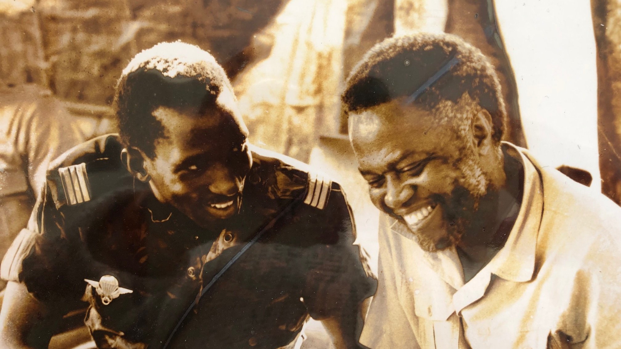 L'image présente deux hommes assis côte à côte, souriant et en train de discuter. L'un d'eux porte un uniforme militaire, tandis que l'autre est habillé d'une chemise claire. Leurs visages expriment la joie et la camaraderie. En arrière-plan, on peut distinguer des silhouettes floues de personnes et de la végétation, ce qui suggère un environnement social dynamique. Les teintes de l'image sont sépia, ce qui lui donne un aspect vintage, évoquant une époque passée.