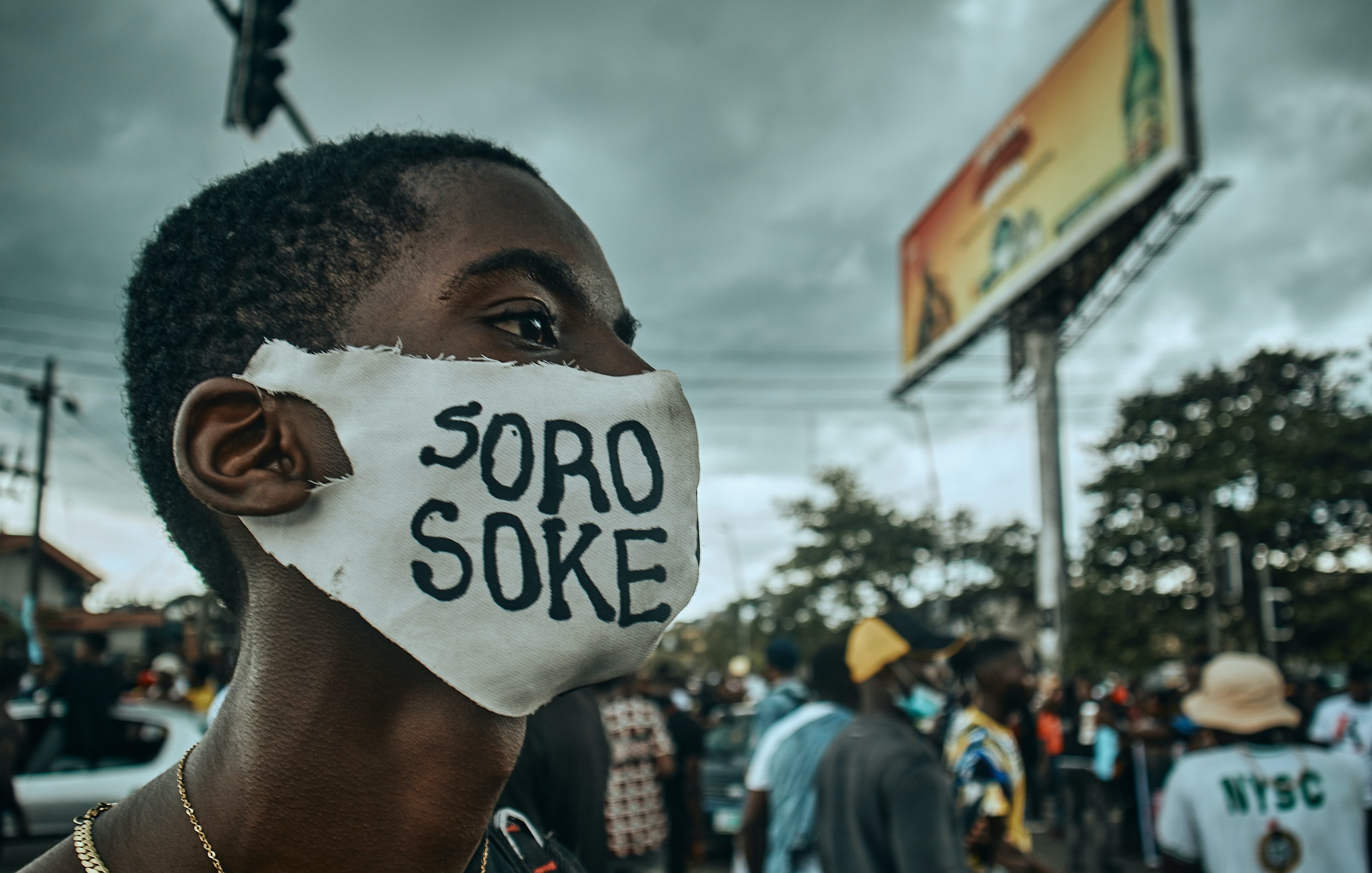 L'image montre une scène de manifestation dans une rue animée. Au premier plan, un jeune homme porte un masque blanc sur lequel est inscrit "SORO SOKE" en lettres noires, un slogan qui évoque la lutte pour les droits et la justice. Son regard est déterminé, et on peut sentir l'énergie de la foule qui l'entoure. À l'arrière-plan, des silhouettes de personnes se déplacent, et des affiches publicitaires peuvent être aperçues. Le ciel est nuageux, ce qui ajoute une atmosphère de tension à la scène. L'ensemble dégage une forte impression d'engagement et de solidarité.