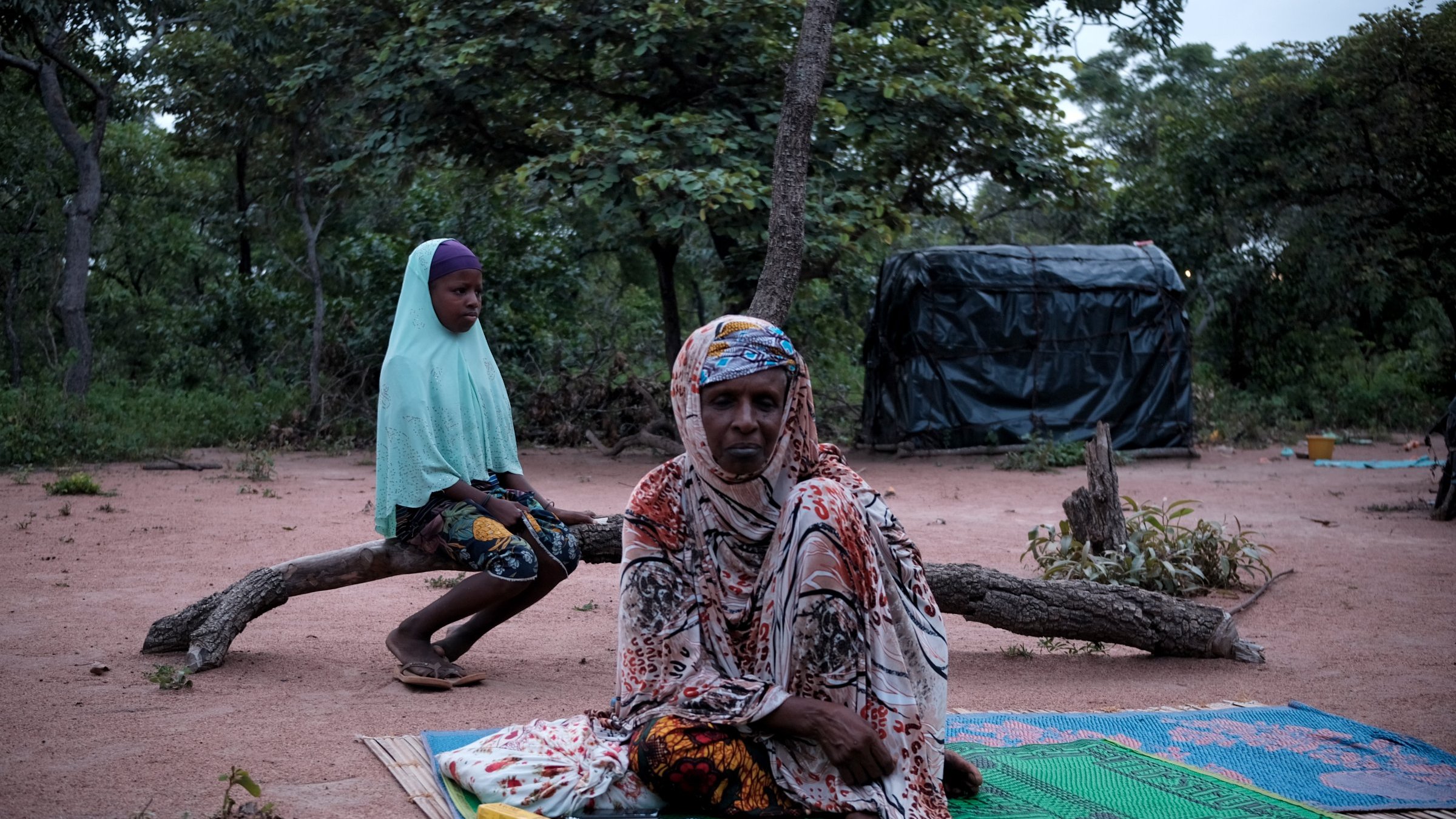 Dans cette image, nous sommes dans un environnement naturel, avec le sol recouvert de terre rouge et entouré d'arbres. À l'avant, une femme est assise sur un tapis, vêtue d'un habit coloré et d'un foulard. Elle a une expression calme. À l'arrière-plan, une jeune fille est assise sur une branche, portant un voile et un vêtement aux motifs colorés. La scène dégage une atmosphère de sérénité et de contemplation, avec la lumière naturelle qui crée une ambiance douce et paisible.