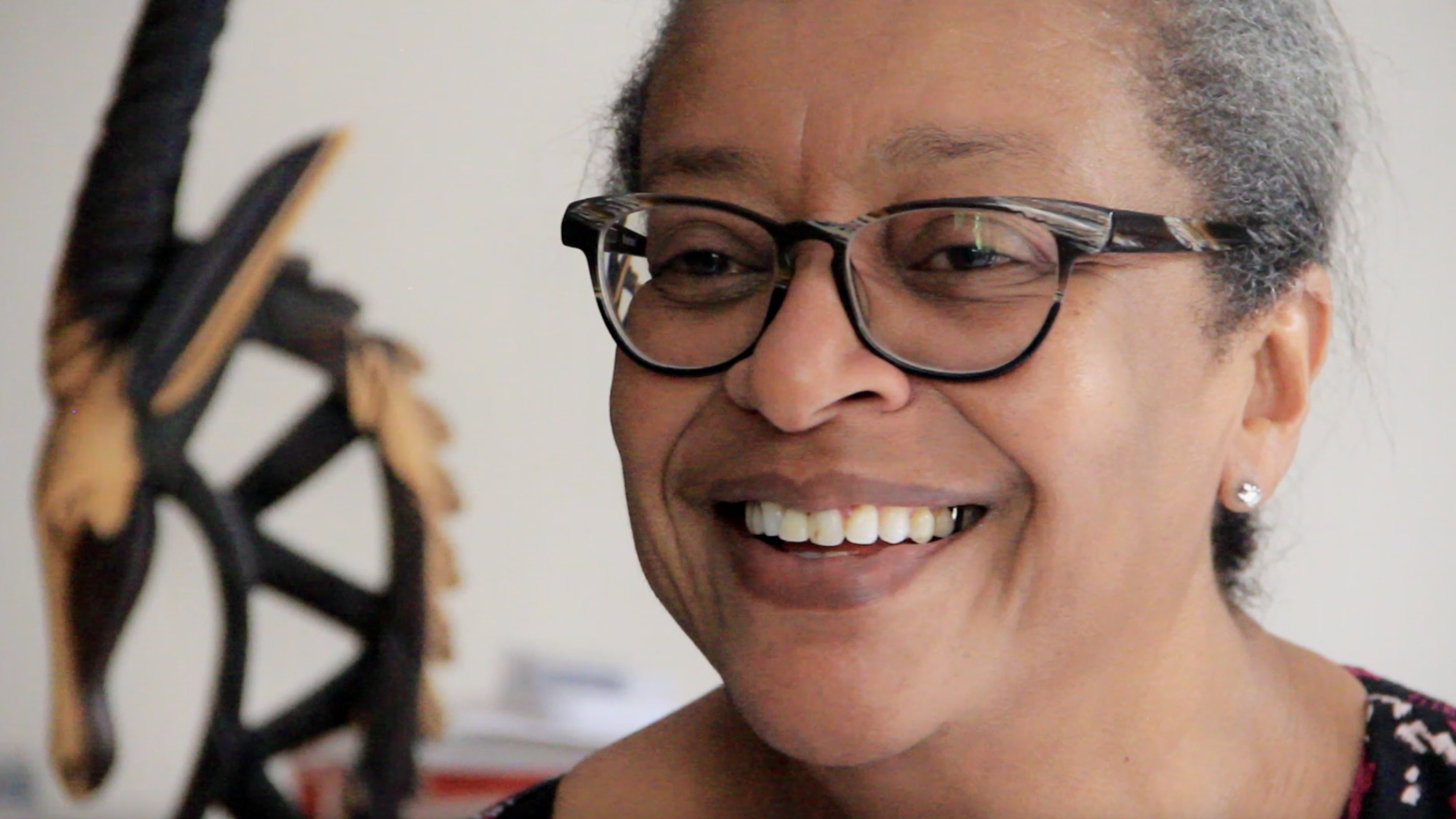 L'image montre une femme souriante, avec des lunettes à monture noire. Son expression est chaleureuse et accueillante. Elle a les cheveux grisonnants, peignés en arrière, et porte des boucles d'oreilles discrètes. En arrière-plan, on aperçoit une sculpture en bois, qui semble représenter un motif stylisé. L'atmosphère est conviviale et intime, suggérant un moment de bonheur ou de partage.