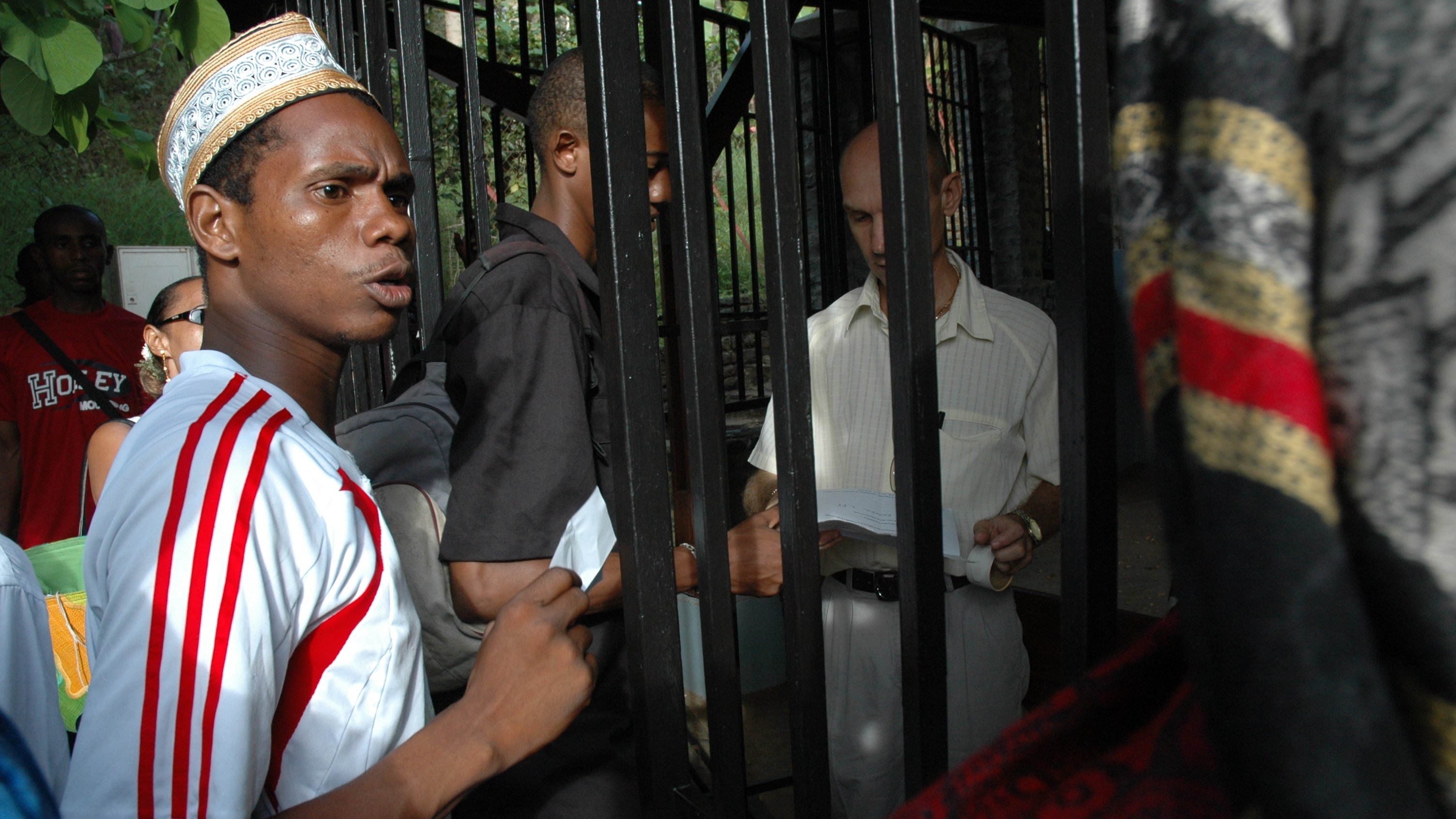 L'image montre un groupe de personnes rassemblées autour d'une structure métallique, probablement une grille ou une clôture. Un jeune homme au premier plan, portant un maillot blanc avec des rayures rouges et un chapeau traditionnel, exprime une certaine agitation ou curiosité. À côté de lui, d'autres personnes, dont un homme en chemise claire, semblent discuter ou échanger des documents. L'environnement est extérieur et verdoyant, suggérant un lieu fréquenté. Les expressions des visages indiquent une interaction dynamique et engageante entre les personnes présentes.