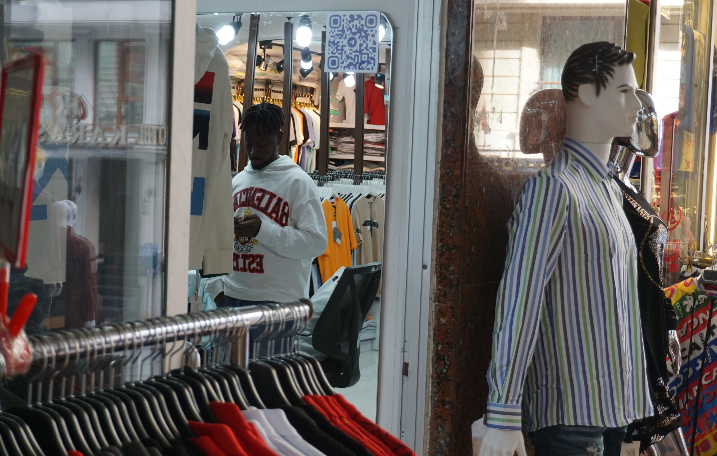 Dans cette image, nous voyons un magasin de vêtements. À l'avant, il y a un mannequin habillé d'une chemise rayée aux couleurs claires, affiché sur une vitrine. Derrière le mannequin, il y a un homme, qui porte un sweat à capuche blanc et semble utiliser son téléphone. Le magasin est bien éclairé, avec des étagères remplies de t-shirts colorés visibles à l'intérieur. Le fond de l'image montre une ambiance animée et des vêtements suspendus.