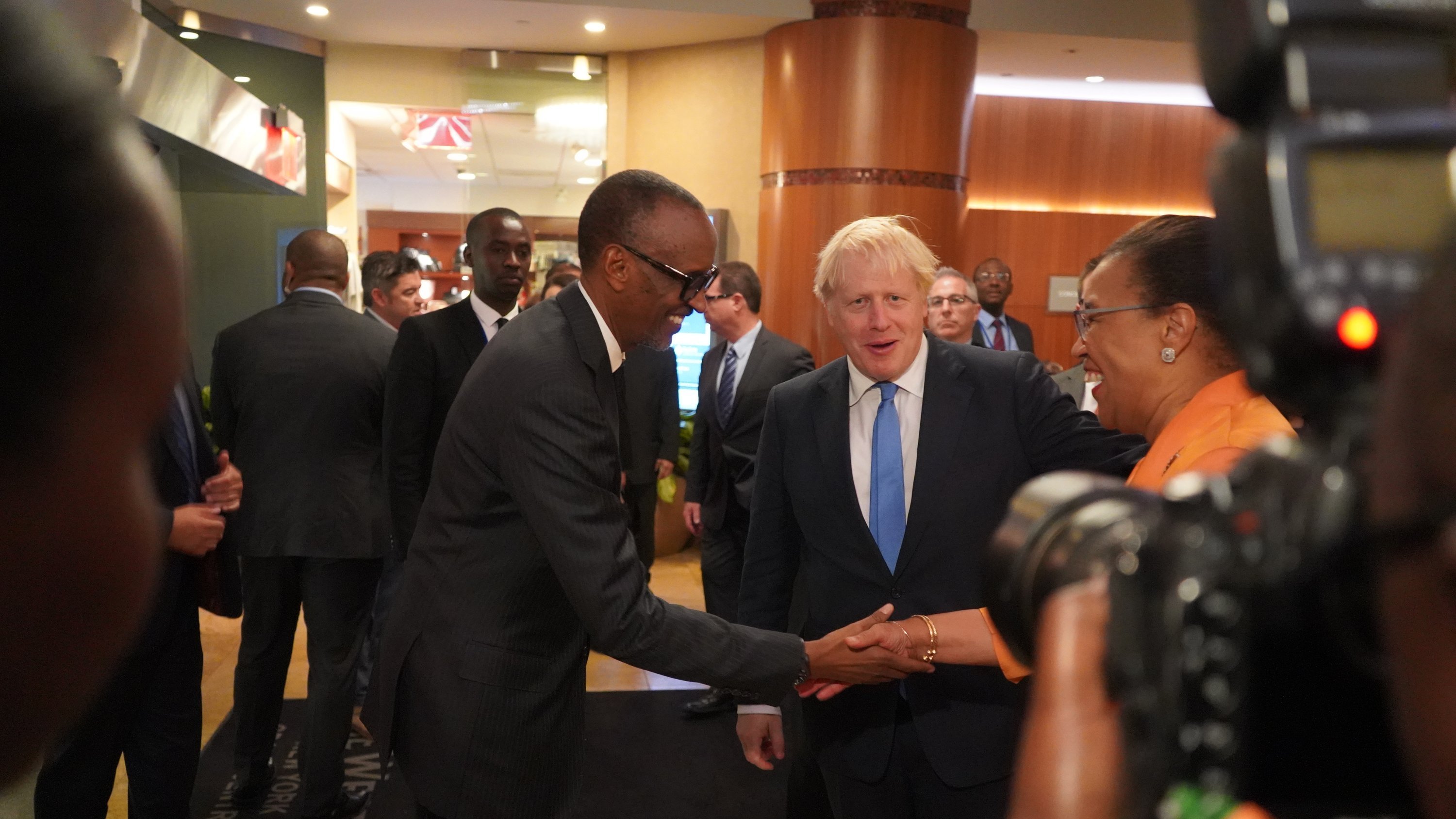 L'image représente une scène dans un hall d'hôtel ou de conférence, où plusieurs personnes sont rassemblées. Au centre, deux hommes se serrent la main : l'un est de grande taille, portant un costume sombre et des lunettes, tandis que l'autre, avec des cheveux blonds, est habillé d'un costume clair. Ils affichent tous deux des sourires amicaux. En arrière-plan, on peut voir plusieurs autres personnes en discussion, ainsi que des détails du décor intérieur, comme des murs aux tons chauds et des éléments de lumière. L'ambiance semble professionnelle et conviviale.