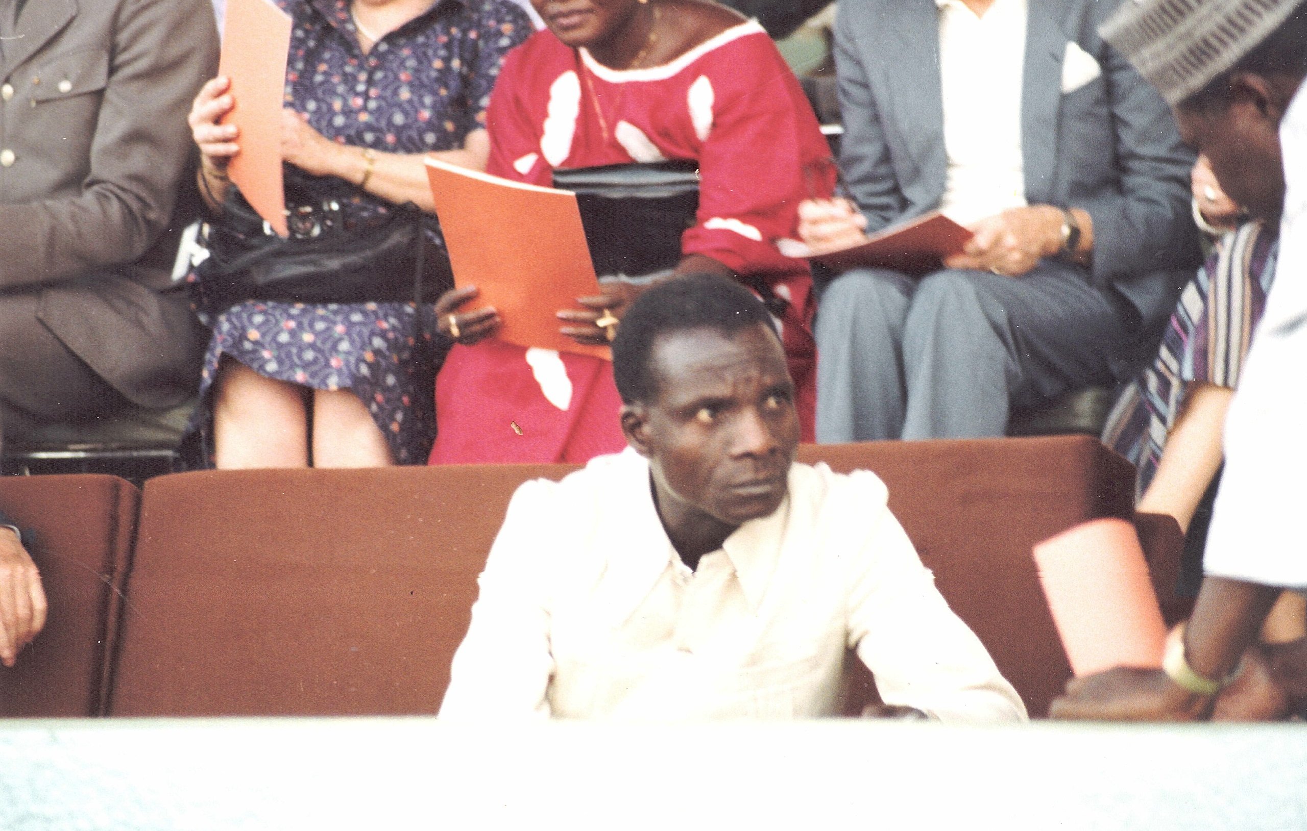 L'image montre une scène d'un événement où un homme en chemise claire est assis au premier plan, semblant sérieux et attentif. À l'arrière-plan, plusieurs personnes discutent, certaines tenant des documents ou des livrets. On aperçoit des vêtements colorés, peut-être d'origine africaine, ce qui ajoute une dimension culturelle à la scène. L'atmosphère générale évoque un rassemblement formel ou une cérémonie.