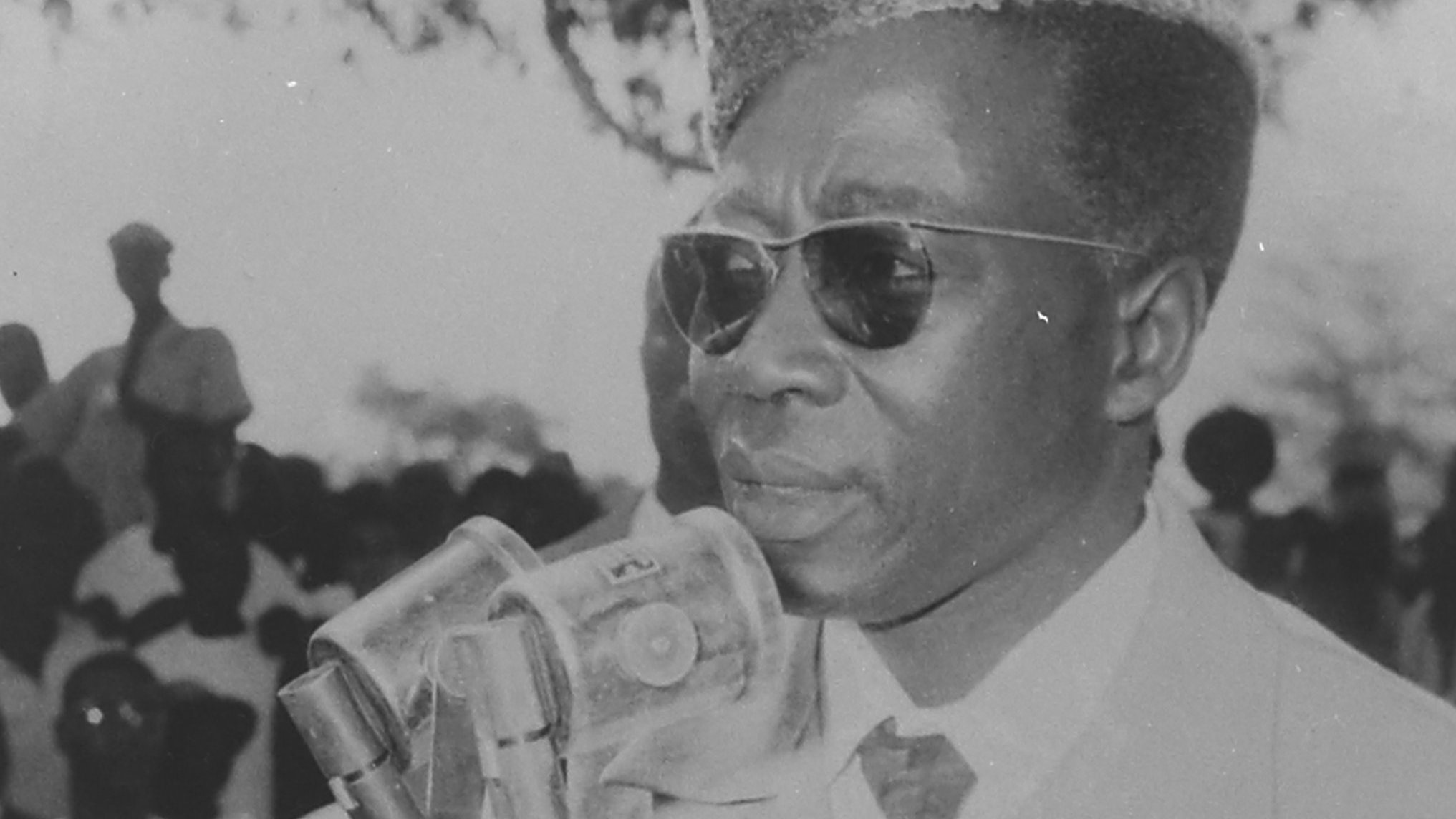 L'image montre un homme debout devant un microphone, s'exprimant clairement. Il porte des lunettes de soleil et a une coiffure distinctive, avec une coupe qui semble bien soignée. Il est vêtu d'un costume clair, ce qui ajoute à son allure sérieuse. En arrière-plan, on peut apercevoir une foule de personnes, dont certaines sont floues, semblant écouter attentivement. L'atmosphère de la scène évoque un moment important, peut-être un discours ou une déclaration publique.