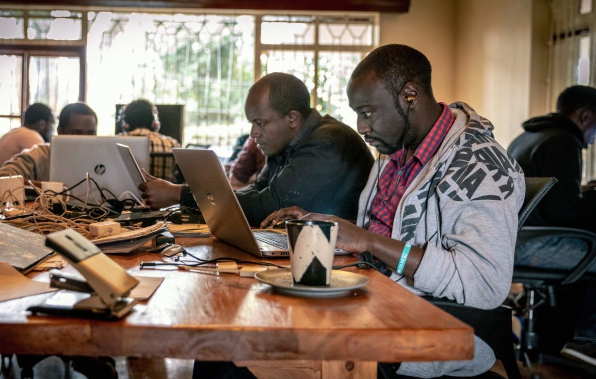 Dans cette image, on voit un espace de travail collaboratif. Plusieurs personnes sont assises autour d'une grande table en bois. Chaque personne utilise un ordinateur portable, principalement des MacBook. Il y a des câbles qui traînent sur la table, ajoutant à l'ambiance de travail intensif. À droite, un homme porte un sweat à capuche gris et écoute des écouteurs, concentré sur son écran. Sur la table, on aperçoit une tasse, peut-être de café, ainsi que quelques feuilles. Les murs sont clairs et de grandes fenêtres laissent entrer la lumière naturelle, créant une atmosphère chaleureuse et productive. En arrière-plan, d'autres personnes sont visibles, toutes absorbées dans leur travail.