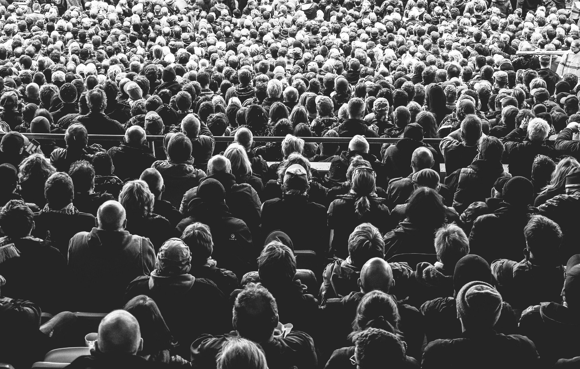 L'image montre une immense foule de personnes rassemblées dans un espace, probablement lors d'un événement ou d'un concert. Les individus sont vus de dos, et leurs têtes sont variées en termes de coiffure et de couleur de cheveux. La majorité des personnes semblent concentrées et immergées dans l'événement, créant une atmosphère collective. Les jeux d'ombres et de lumières donnent à l'image un aspect noir et blanc, accentuant la densité de la foule et le sentiment d'unité parmi les spectateurs. On peut presque sentir l'énergie vibrante qui se dégage de ce groupe immense.