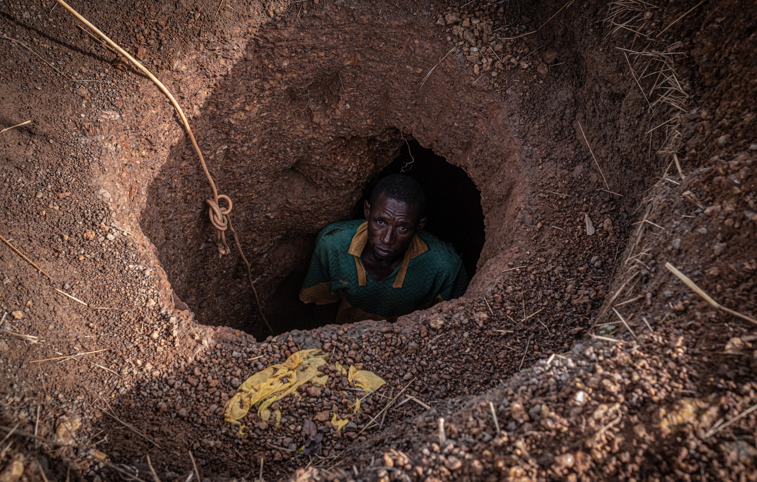 Dans cette image, nous voyons un homme qui se tient au fond d'un trou creusé dans le sol. Le trou a une forme ronde, et les parois sont en terre brune, légèrement humides et irrégulières. L'homme, vêtu d'un maillot vert avec un col jaune, regarde vers le haut avec une expression sérieuse. Son visage est en partie dans l'ombre, mais son regard est visible. Autour du trou, on peut apercevoir des gravats et des brindilles qui ajoutent un aspect naturel et brut à l'environnement. La scène évoque un sentiment d'isolement et de travail ardu.