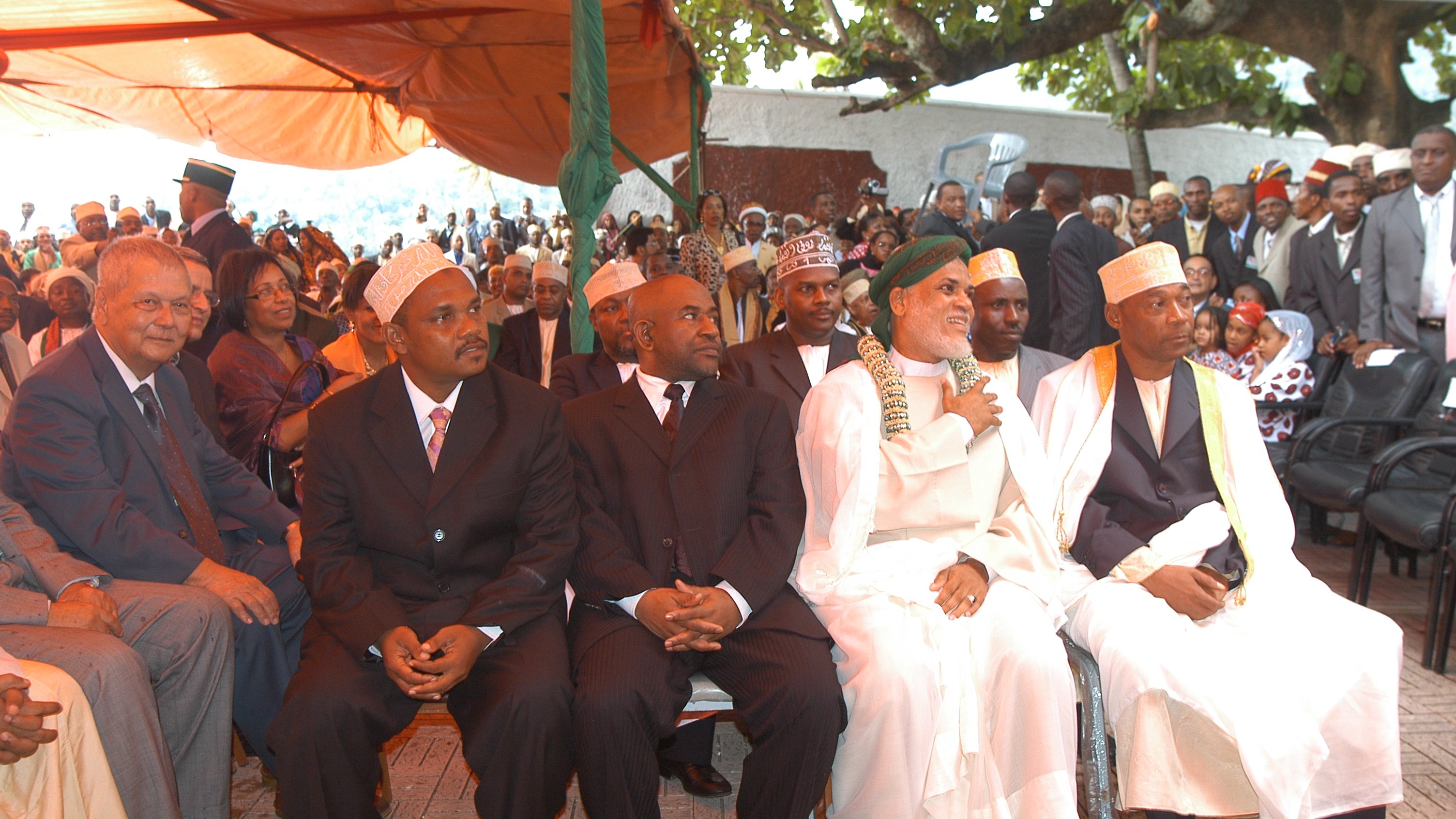 L'image montre un groupe d'hommes assis côte à côte lors d'un événement en plein air. Au premier plan, quatre hommes bien habillés portent des costumes sombres et des chapeaux traditionnels. Le premier homme à gauche a des lunettes et affiche une expression sérieuse. Le deuxième, au milieu, est habillé en noir et semble concentré. Le troisième, portant une robe traditionnelle blanche et un turban, sourit et semble être en conversation. Le quatrième homme, également en tenue traditionnelle, regarde droit devant lui. En arrière-plan, on peut apercevoir une foule de personnes rassemblées, avec des arbres et des tentes fournissant de l'ombre. L'ambiance paraît formelle et respectueuse, suggérant que ce moment est empreint de signification culturelle ou de cérémonie importante.