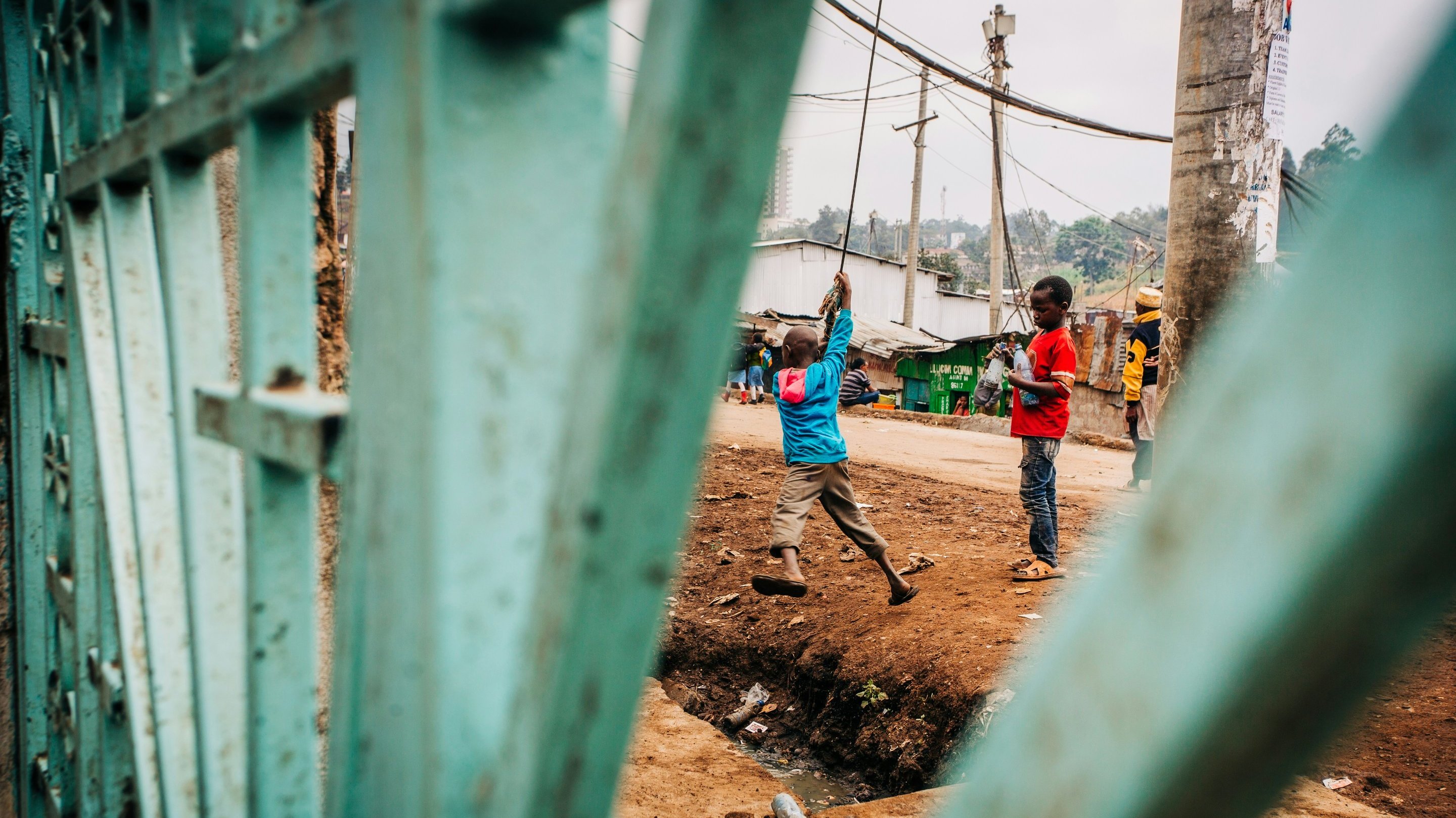L'image présente une scène de la vie quotidienne dans un environnement urbain. Au premier plan, on aperçoit des barres d'une grille verte, qui encadrent la vue. À l'intérieur, deux enfants jouent près d'un chemin. L'un porte une chemise bleue et tient un objet au-dessus de sa tête, semblant s'amuser. L'autre enfant, aux vêtements rouges, observe ou se prépare à rejoindre le jeu. En arrière-plan, il y a des maisons simples, des poteaux électriques et une ambiance vibrante, typique d'un quartier animé. On sent une atmosphère de camaraderie et d'énergie enfantine. Le terrain est boueux et légèrement en pente.