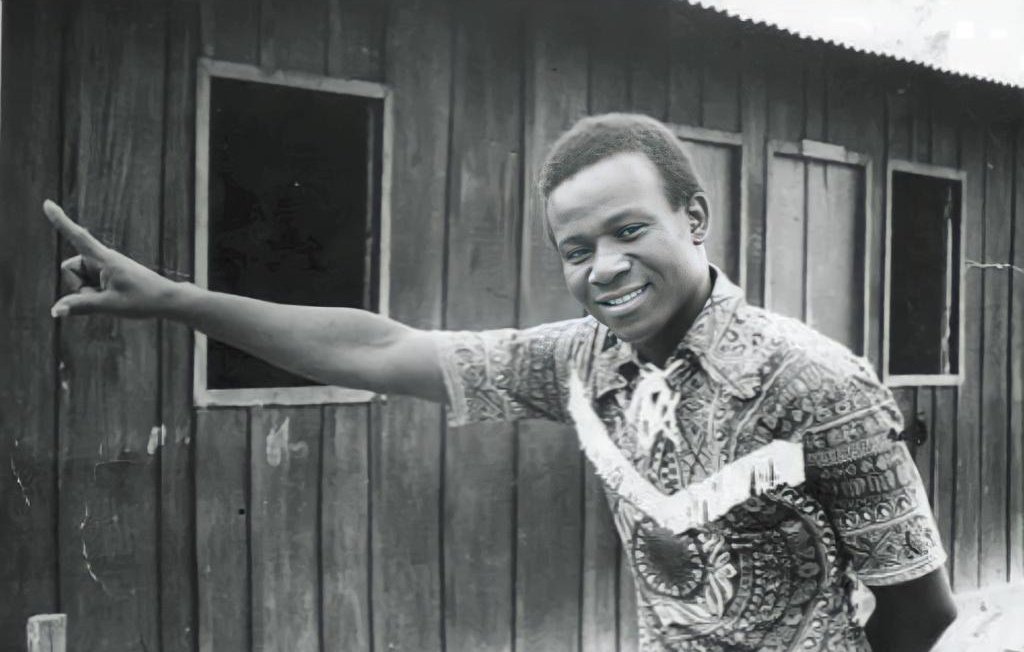 L'image montre un homme debout à côté d'une maison en bois. Il porte une chemise colorée au motif complexe, avec des nuances de marron et de beige. Son sourire est amical et engageant. L'homme pointe une main vers un mur en bois, suggérant une direction ou attirant l'attention sur quelque chose. En arrière-plan, on aperçoit des fenêtres sombres qui ajoutent une dimension de profondeur à la scène. L'atmosphère semble à la fois chaleureuse et authentique, évoquant un moment de convivialité.