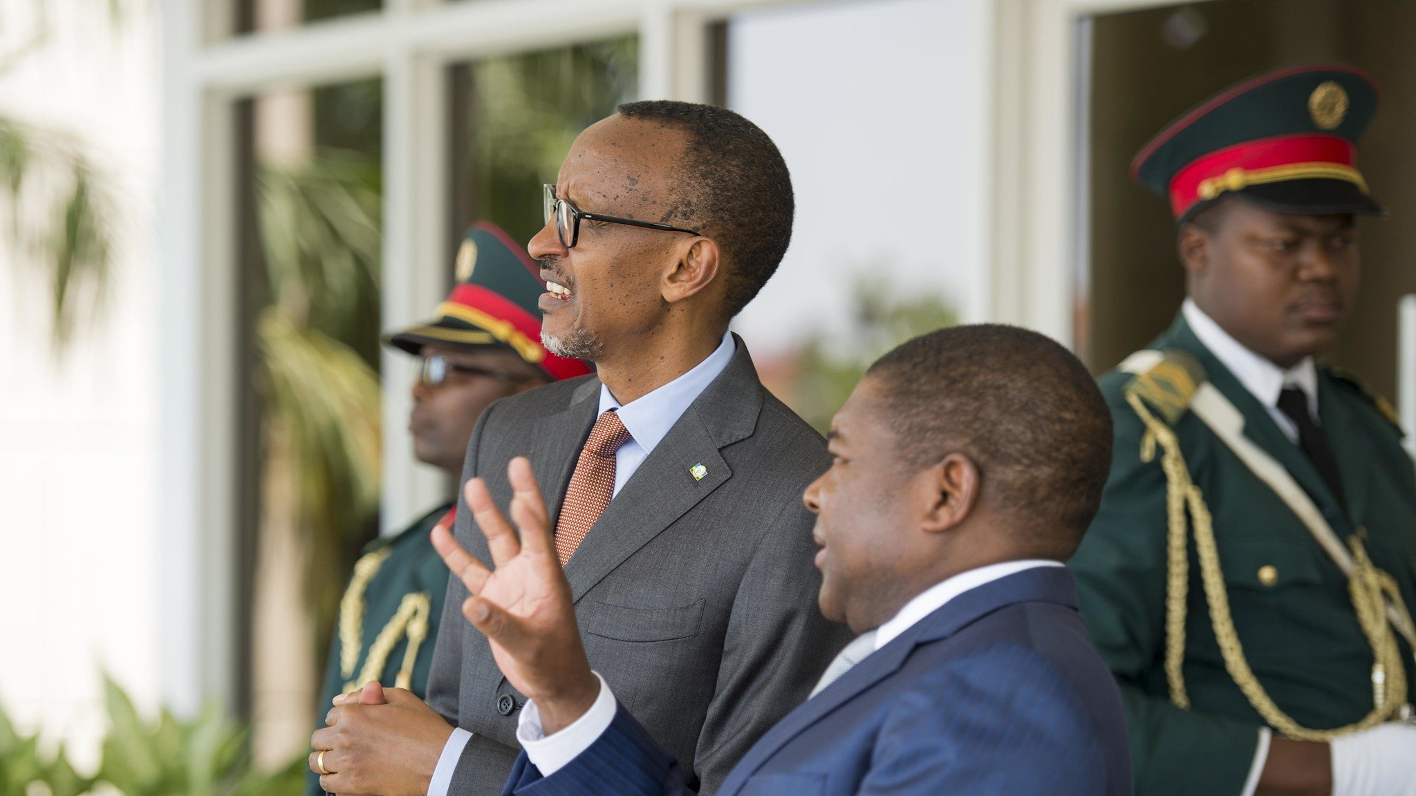 Sur cette image, nous voyons deux hommes en costume dialoguant dans un cadre officiel. L'homme à gauche, avec des lunettes et un sourire, semble être en train d'écouter attentivement. À sa droite, un homme aux cheveux courts gesticule avec les mains, soulignant un point dans la conversation. En arrière-plan, deux gardes en uniforme militaire se tiennent, assurant une ambiance de sécurité. On peut également percevoir des éléments de verdure, suggérant un environnement extérieur agréable.