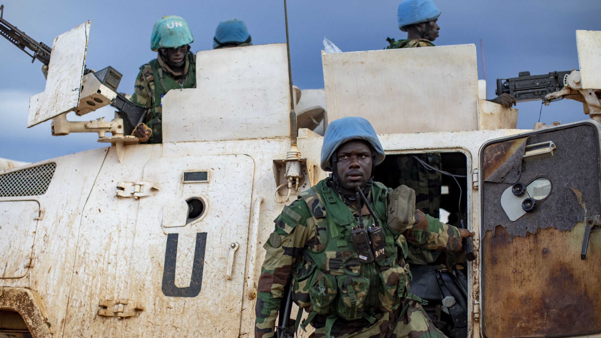 L'image montre un groupe de soldats en uniforme, portant des casques bleus emblématiques des Casques bleus de l'ONU. Ils sont près d'un véhicule militaire, sale et marqué par la boue, ce qui indique des conditions difficiles. Le soldat en premier plan, avec un regard déterminé, semble descendre du véhicule. À l'arrière, d'autres soldats se tiennent, prêts à intervenir. Le ciel est nuageux, ajoutant une ambiance sombre et sérieuse à la scène, qui évoque une mission de maintien de la paix dans un environnement hostile.