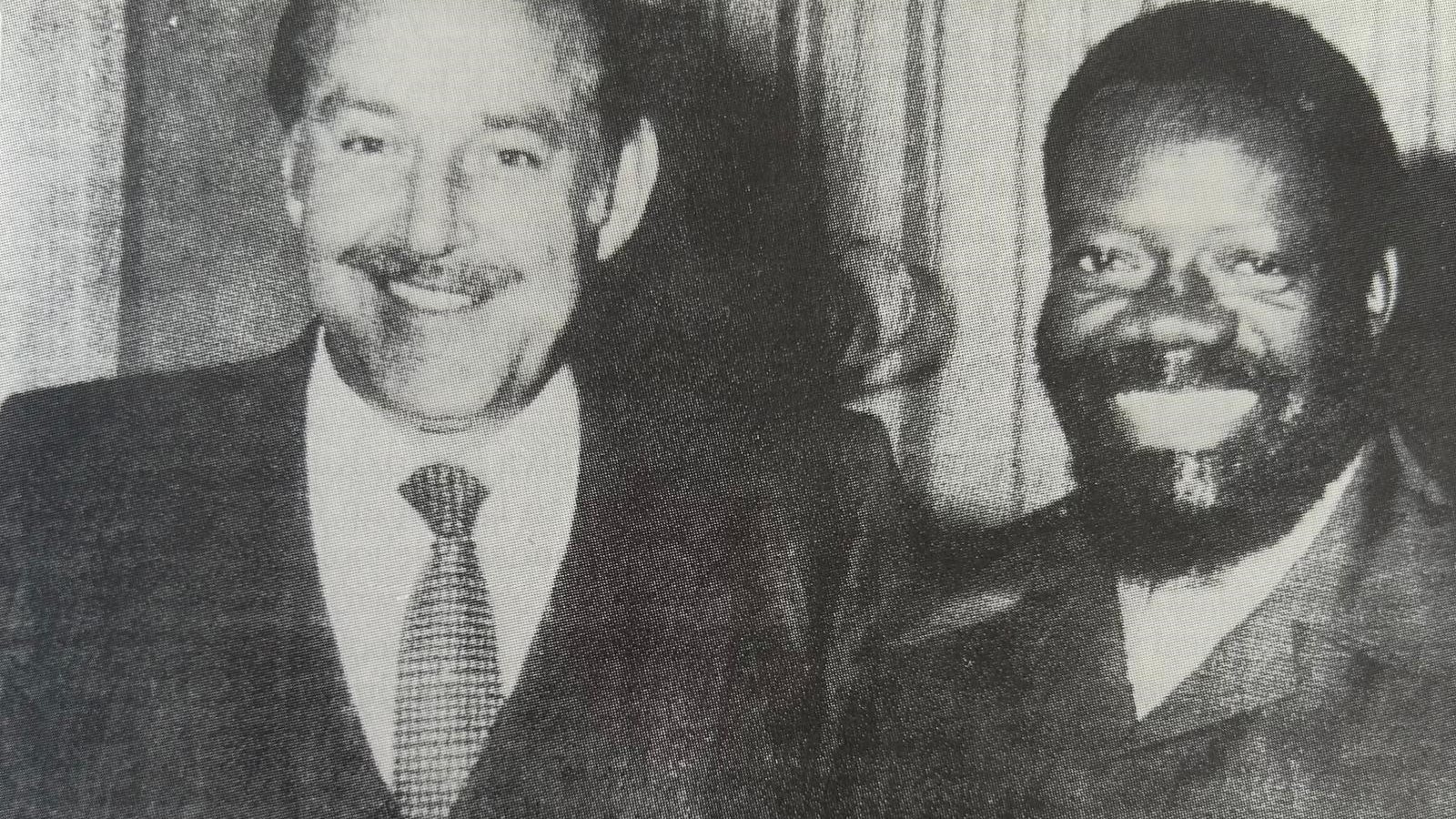 L'image montre deux hommes se tenant côte à côte, souriant largement. Le premier homme, à gauche, porte un costume sombre avec une cravate. Il a des cheveux grisonnants, une moustache bien dessinée et une expression amicale. Le deuxième homme, à droite, est habillé d'une veste de style militaire et a une barbe bien fournie. Il a les cheveux frisés et sourit également, ce qui donne une atmosphère chaleureuse à la photo. Les deux tiennent des verres à la main, suggérant une occasion conviviale, peut-être une célébration ou un événement. L'arrière-plan est flou, mettant l'accent sur les deux hommes.