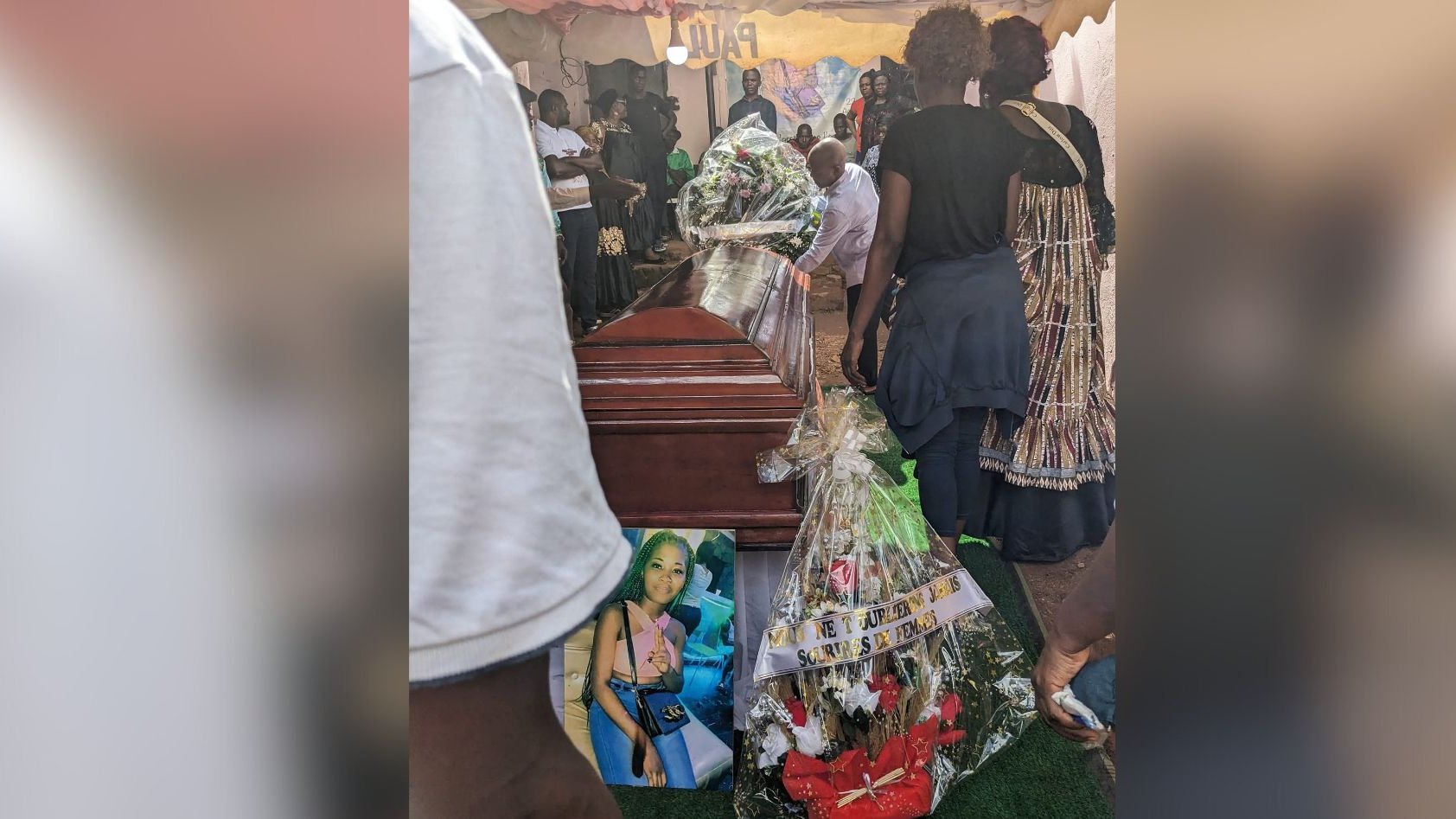 L'image montre un moment poignant d'un enterrement. Au centre, un cercueil en bois brun, sur lequel sont déposés des bouquets de fleurs. À côté du cercueil, une grande photo d'une femme est accrochée, entourée de décorations florales et de bandes de tissu. Des personnes, visiblement en deuil, se rassemblent autour, certaines portant des vêtements sombres. L'ambiance est sombre mais respectueuse, marquée par la tristesse et l'hommage à la personne décédée. L'arrière-plan est flou, mais on peut deviner une tente qui abrite cette cérémonie. L'ensemble évoque des sentiments de perte et de commémoration.