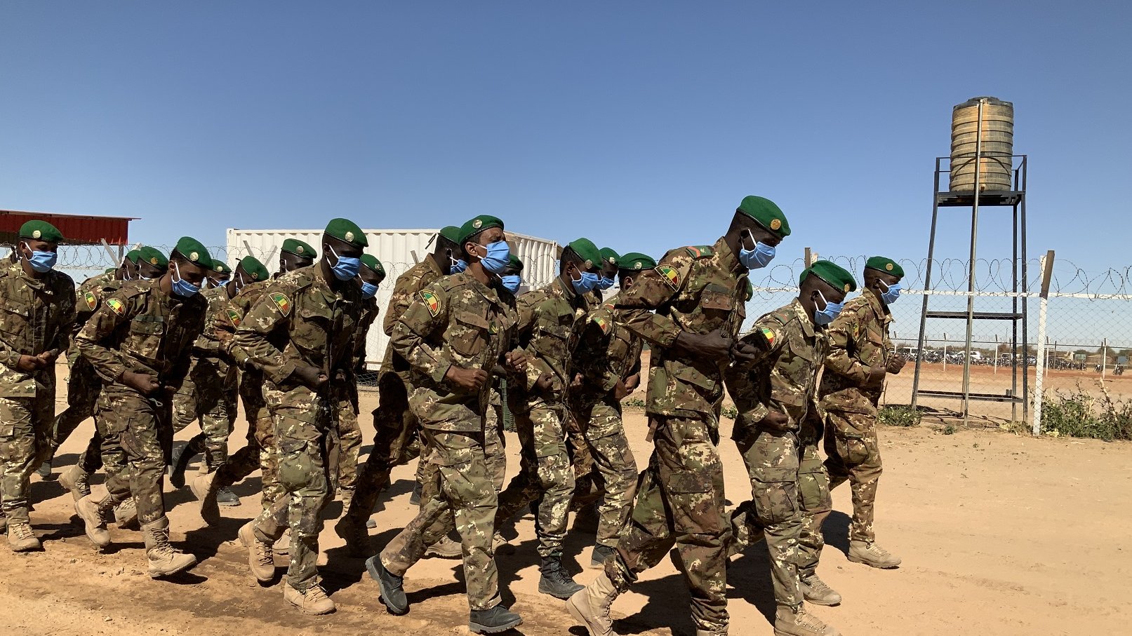Dans cette image, un groupe de soldats est en train de marcher en formation sur un chemin poussiéreux. Ils portent des uniformes militaires verts camouflés et des casquettes vertes. La majorité d'entre eux portent également des masques bleus, ce qui suggère une mesure de protection sanitaire. En arrière-plan, on peut distinguer des conteneurs et une tour d'eau. Le ciel est dégagé, avec une couleur bleue intense, créant un contraste avec la terre orange et les silhouettes des soldats. L'atmosphère évoque un moment de discipline et d'organisation dans un environnement militaire.