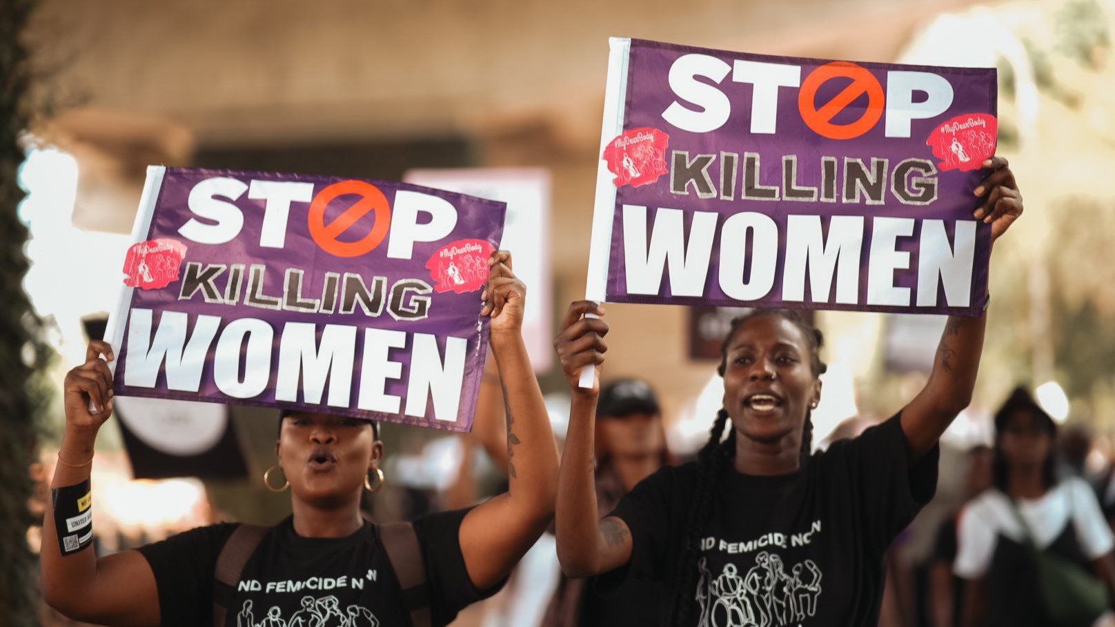 L'image montre deux femmes participante à une manifestation. Elles tiennent chacune une grande pancarte. Les pancartes portent le message "STOP KILLING WOMEN" en lettres capitales de couleur blanche, avec un symbole barré en rouge. Les femmes ont des expressions déterminées et sont vêtues de t-shirts noirs. L'arrière-plan suggère une ambiance de mobilisation avec d'autres personnes qui marchent et participent à l'événement. L'atmosphère est chargée d'émotion et de solidarité.