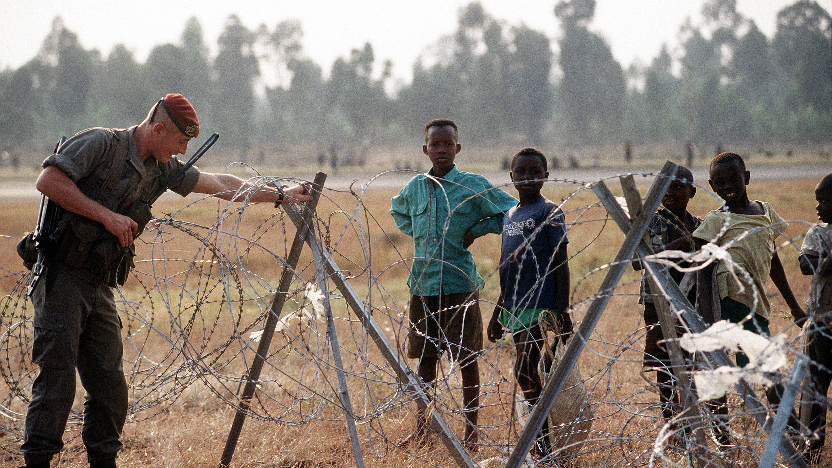 L'image présente un soldat en uniforme, concentré sur un fil barbelé, qui semble inspecter la barrière. Il est à proximité d'un groupe d'enfants qui l'observent. Les enfants, de différentes couleurs de vêtements, affichent des expressions variées, allant de la curiosité à l'inquiétude. L'arrière-plan montre un paysage ouvert, avec des essences d'arbres floues et des herbes sèches au sol. L'atmosphère dégage un mélange d'autorité et d'innocence, avec le soldat s'occupant de la sécurité, tandis que les enfants représentent l'innocence de l'enfance dans un contexte potentiellement difficile.