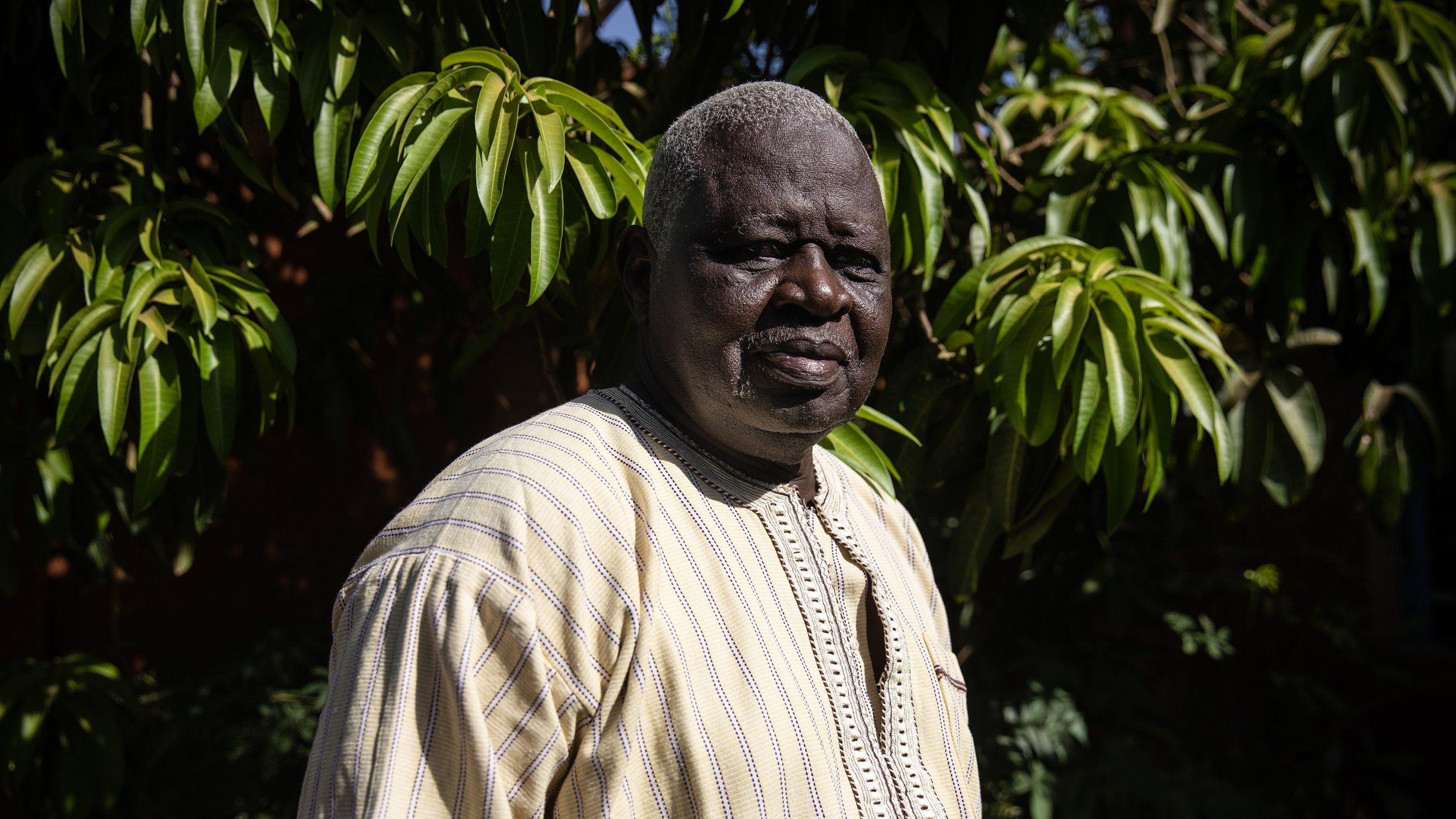 L'image présente un homme debout dans un environnement extérieur verdoyant. Il porte une tunique ample de couleur claire, ornée de motifs discrets, qui témoigne d'un style traditionnel. Son visage est marqué par des traits qui dégagent une certaine sagesse et sérénité. Les cheveux de l'homme sont grisonnants, ajoutant à son apparence mature. En arrière-plan, on aperçoit un feuillage dense et luxuriant, créant un contraste avec sa tenue. La lumière naturelle met en valeur les détails de son visage et de son vêtement, tout en évoquant une atmosphère paisible et accueillante.