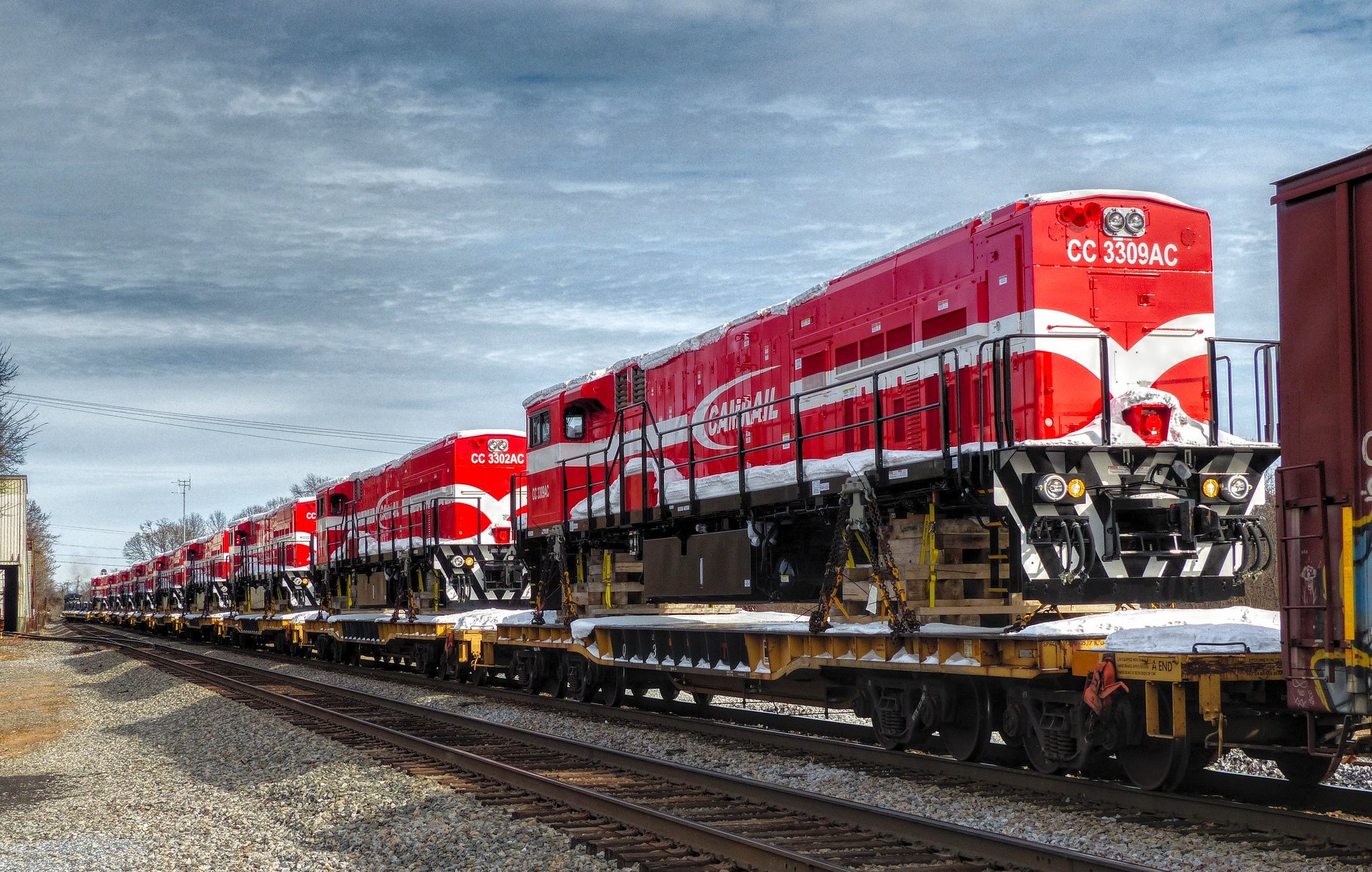 L'image montre une série de locomotives de train d'un rouge vif, alignées sur une flatcar (wagon plat) le long des rails. Les locomotives ont un design distinctif avec des motifs blancs et une grande inscription sur le côté. Le ciel est partiellement nuageux, créant un contraste avec la couleur vive des locomotives. Le cadre suggère un environnement industriel, avec des rails de chemin de fer visibles au premier plan et un décor sobre en arrière-plan. L'atmosphère est calme, typique d'une zone où le train est stationné.