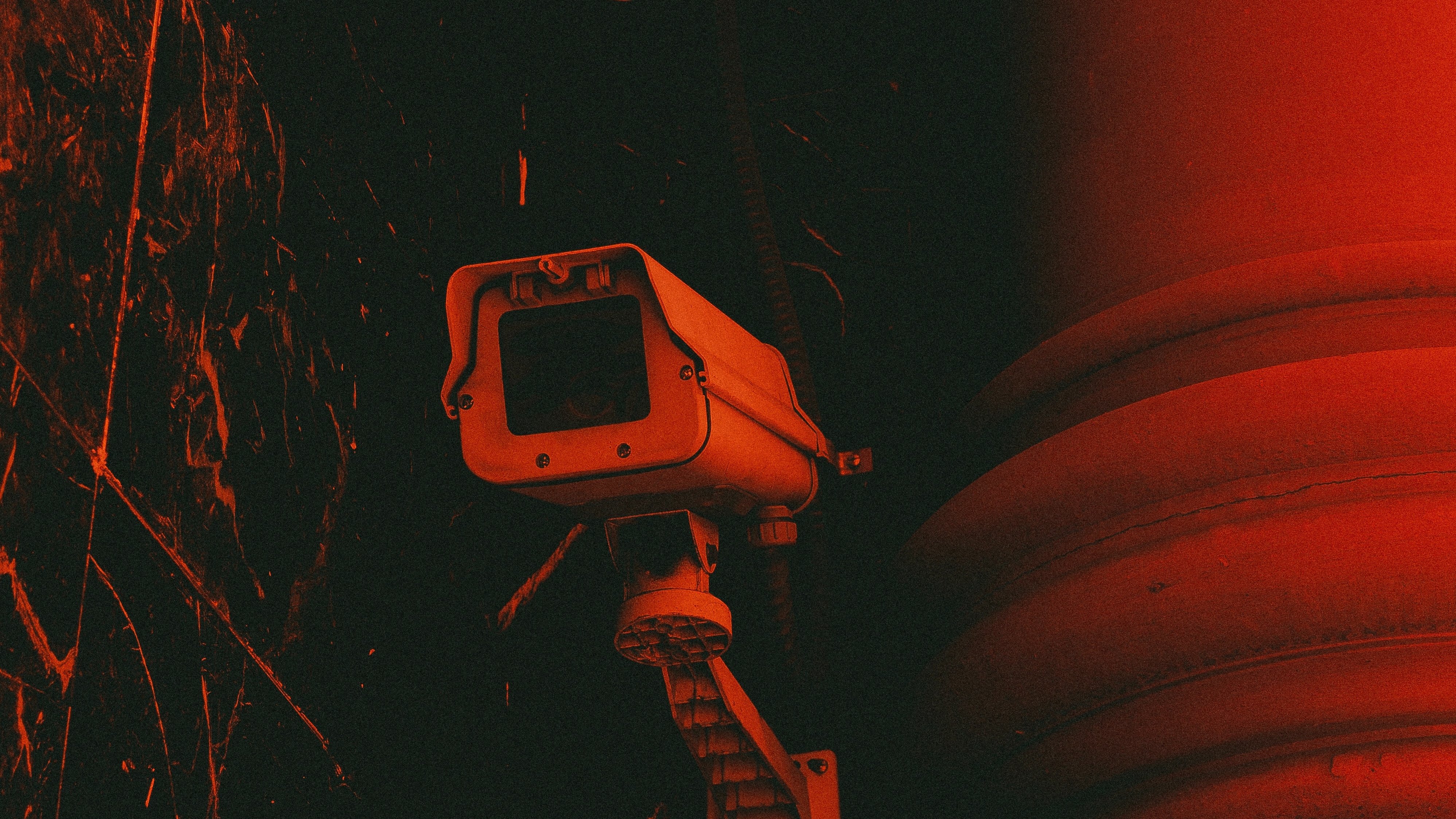 L'image représente une caméra de surveillance fixée à un mur. La caméra, avec son boîtier rectangulaire et son objectif, est peinte en gris clair. Elle est montée sur un support qui lui permet de pivoter, capturant ainsi une vaste zone de surveillance. L'arrière-plan est d'un rouge intense, créant une atmosphère sombre et légèrement inquiétante. Des textures sombres et des motifs abstraits se dessinent autour, ajoutant à l'effet dramatique de l'image. Cette combinaison de couleurs et de formes suggère une ambiance de vigilance et de sécurité.