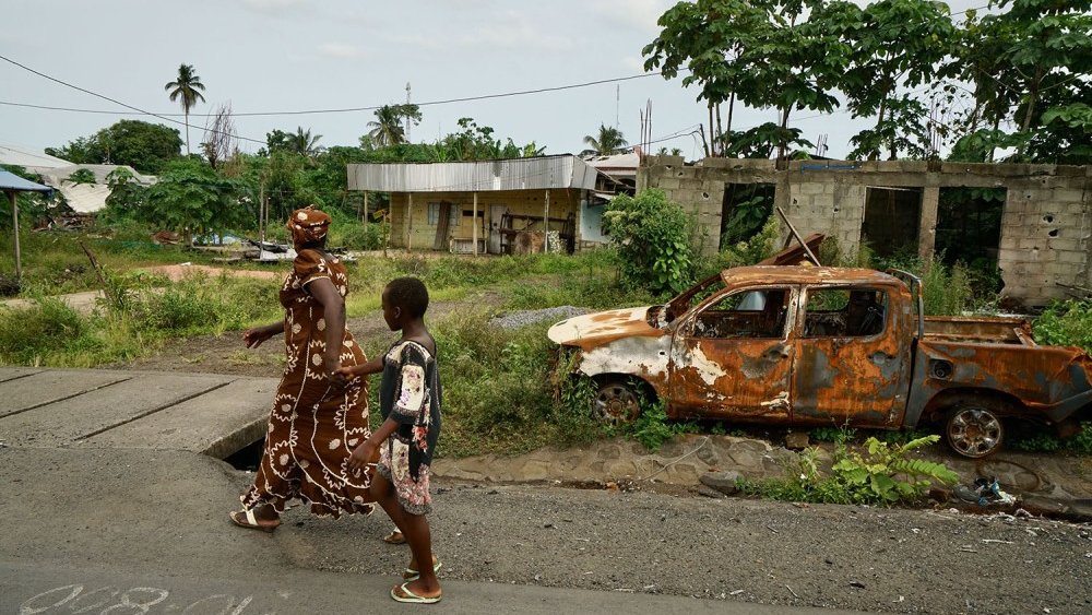 L'image montre une scène d'une rue dans un environnement rural ou semi-urbain. Sur le trottoir, une femme vêtue d'une longue robe ornée de motifs blancs marche aux côtés d'un jeune garçon. Ils avancent côte à côte, probablement en direction d'un endroit précis. En arrière-plan, on peut voir un paysage verdoyant avec des arbres, ainsi que des bâtiments partiellement construits, évoquant une ambiance de quartier en développement. À droite, un vieux véhicule, rouillé et en piteux état, est abandonné sur le côté de la route, entouré de végétation. Le ciel est nuageux, suggérant qu'il pourrait faire chaud et humide. L'ensemble dégage une atmosphère à la fois paisible et un peu nostalgique.