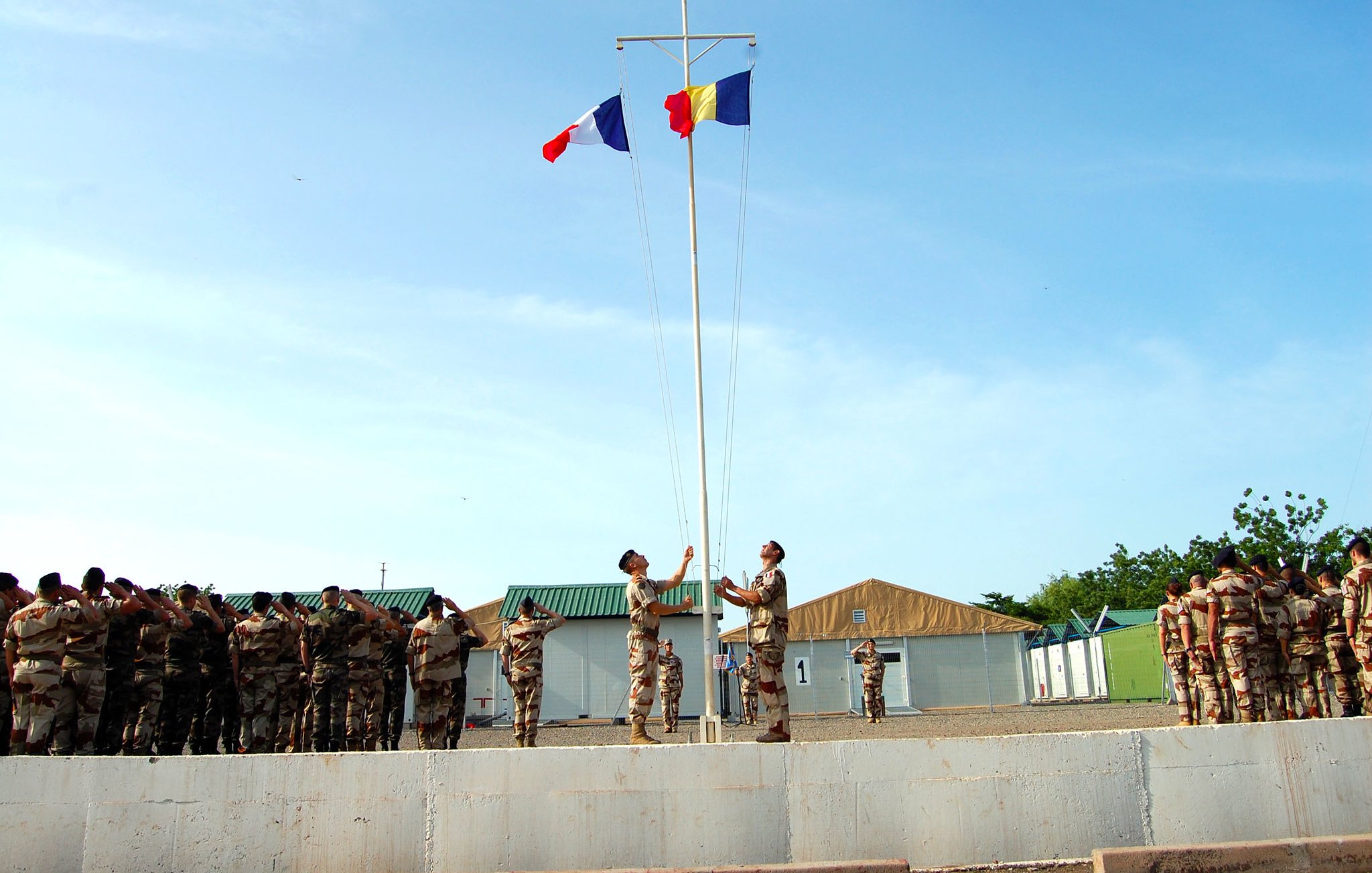 Cette image montre une cérémonie de lever de drapeaux dans un environnement militaire. Au premier plan, deux soldats se tiennent près d'un mât, où ils sont en train de hisser les drapeaux français et roumain. Ils sont parfaitement alignés et semblent respectueux. En arrière-plan, un groupe de soldats en uniforme assiste à la cérémonie, formant une ligne. L'environnement est clair et ensoleillé, avec des bâtiments militaires simples et une légère brise qui pourrait faire flotter les drapeaux. L'atmosphère est solennelle et empreinte de respect.