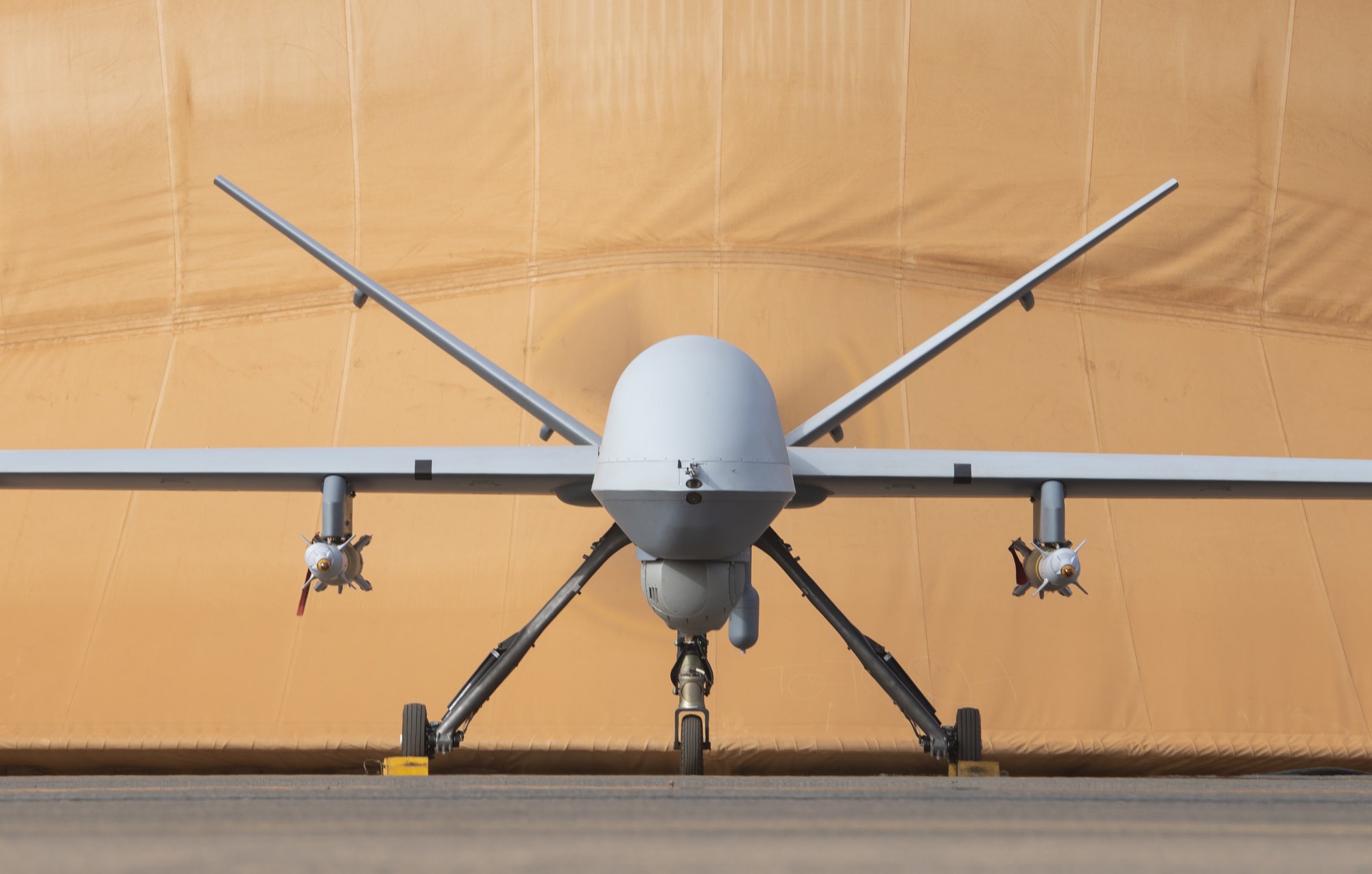 L'image montre un drone militaire de type MQ-9 Reaper. Il est positionné de face, avec le corps fuselé et l'aile légèrement étendue. À chaque côté du fuselage, on peut voir des réservoirs ou des munitions. En arrière-plan, il y a une toile beige qui semble un hangar ou une protection, lui donnant un aspect de préparation à un vol. Le sol est en béton, créant un environnement d'aérodrome. L'ensemble est impressionnant, évoquant la technologie aéronautique moderne.