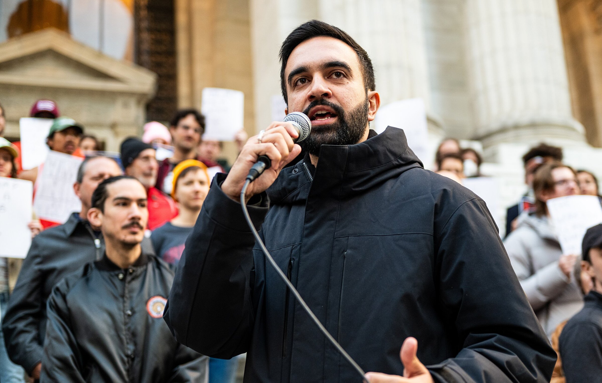 L'image montre un homme debout en avant-plan, tenant un microphone dans sa main et s'adressant à une foule. Il porte une veste noire et a une expression déterminée sur son visage. En arrière-plan, on aperçoit un groupe de personnes, certaines tenant des affiches. L'environnement semble être une place publique, avec des bâtiments importants en arrière-plan. L'atmosphère dégage une impression de mobilisation et d'engagement collectif.
