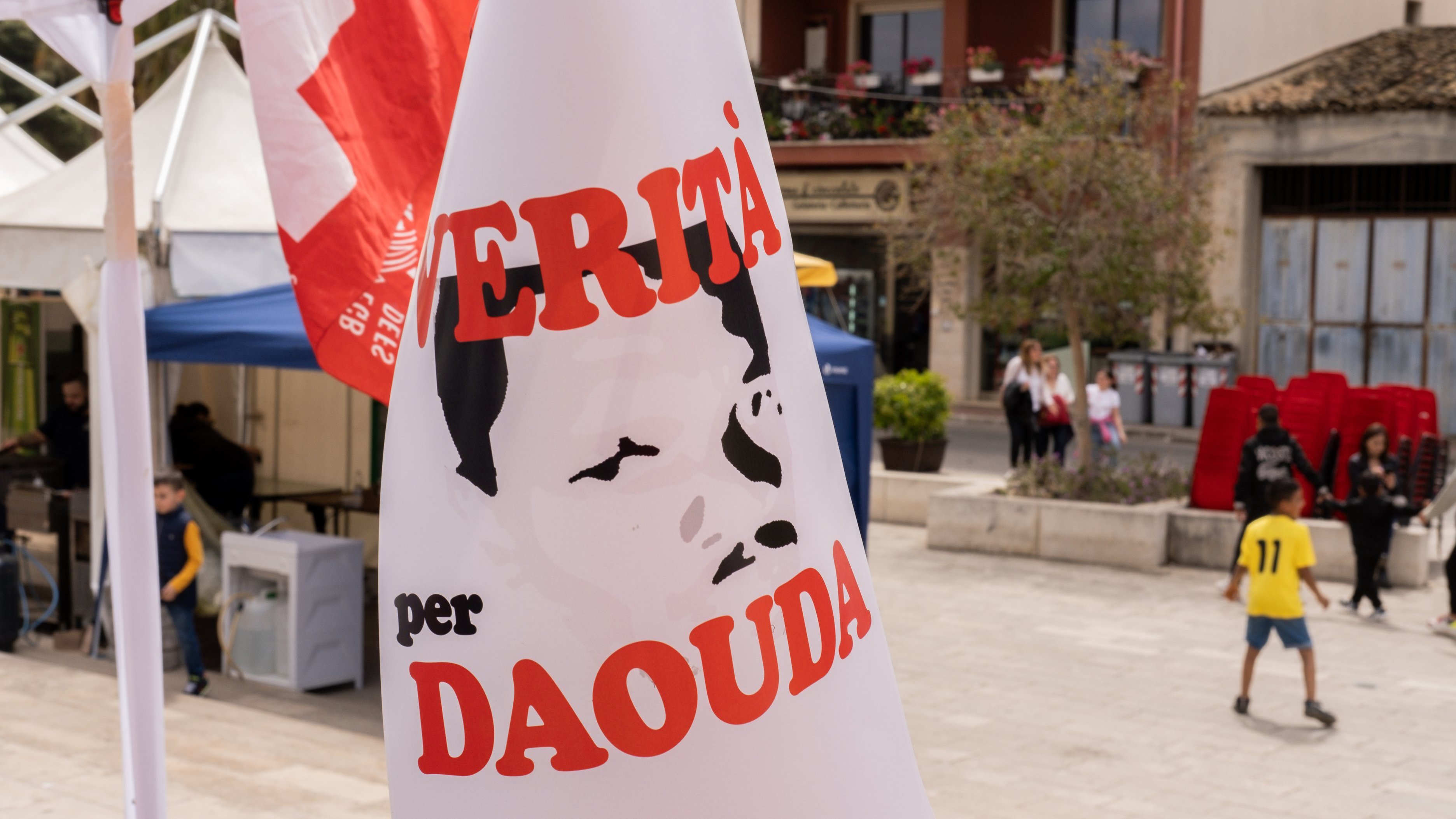 L'image présente une scène en plein air, probablement lors d'un événement ou d'une manifestation. Un panneau est palpable, affichant un portrait stéréotypé d'une personne, dont les traits sont simplifiés et en noir et blanc. Le texte en rouge indique "VERITÀ per DAOUDA", ce qui signifie "Vérité pour Daouda". En arrière-plan, on aperçoit des bâtiments avec des balcons et des tentes blanches. L'atmosphère semble engagée, avec un mélange de couleurs vives des drapeaux et des vêtements des personnes présentes. Le son des conversations et des rires pourrait également emplir l'air, créant une ambiance dynamique et communautaire.