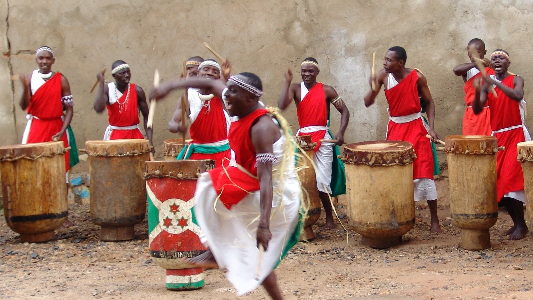 L'image représente un groupe de percussionnistes en pleine performance. Ils portent des vêtements traditionnels, principalement rouges, avec des ceintures vertes. Au centre, un homme se distingue en sautant avec énergie, créant un mouvement dynamique. Les autres membres du groupe, en arrière-plan, jouent des tambours en bois de forme cylindrique. L'atmosphère est festive et rythmée, avec une ambiance de célébration culturelle, où les sons des tambours résonnent fortement. Le sol est en terre, ce qui ajoute un aspect rustique à la scène.