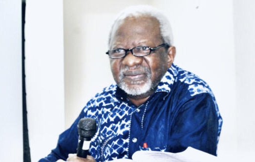 L'image présente un homme assis à une table, tenant un microphone. Il porte des lunettes et a des cheveux gris et bouclés. Son visage exprime une certaine sagesse et réflexion. Il est habillé d'une chemise à motifs, principalement bleue et blanche. Le fond de l'image est neutre, mettant en avant sa présence. On peut sentir une atmosphère de concentration, probablement lors d'un discours ou d'une discussion.
