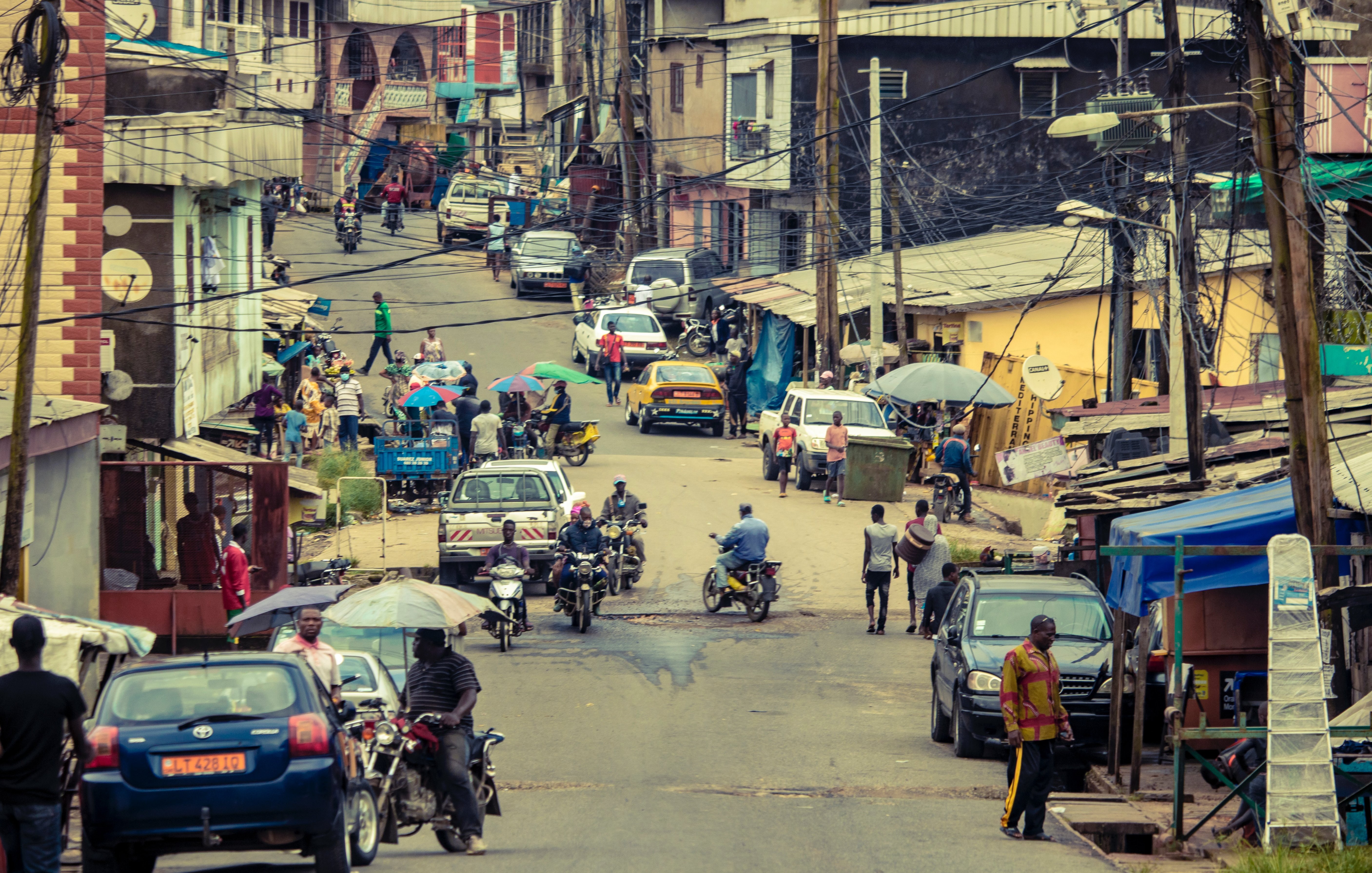 L'image dépeint une rue animée d'une ville. On y voit des bâtiments à plusieurs étages, certains colorés, avec des toits en tôle. La rue est bordée de voitures et de deux roues, et on aperçoit des personnes se déplaçant sur le trottoir. Certaines personnes marchent, d'autres se déplacent à moto. Des parasols colorés sont installés sur le trottoir, offrant de l'ombre, et il y a des fils électriques qui traversent l'espace. L'atmosphère est vivante et dynamique, reflétant le quotidien d'une ville.