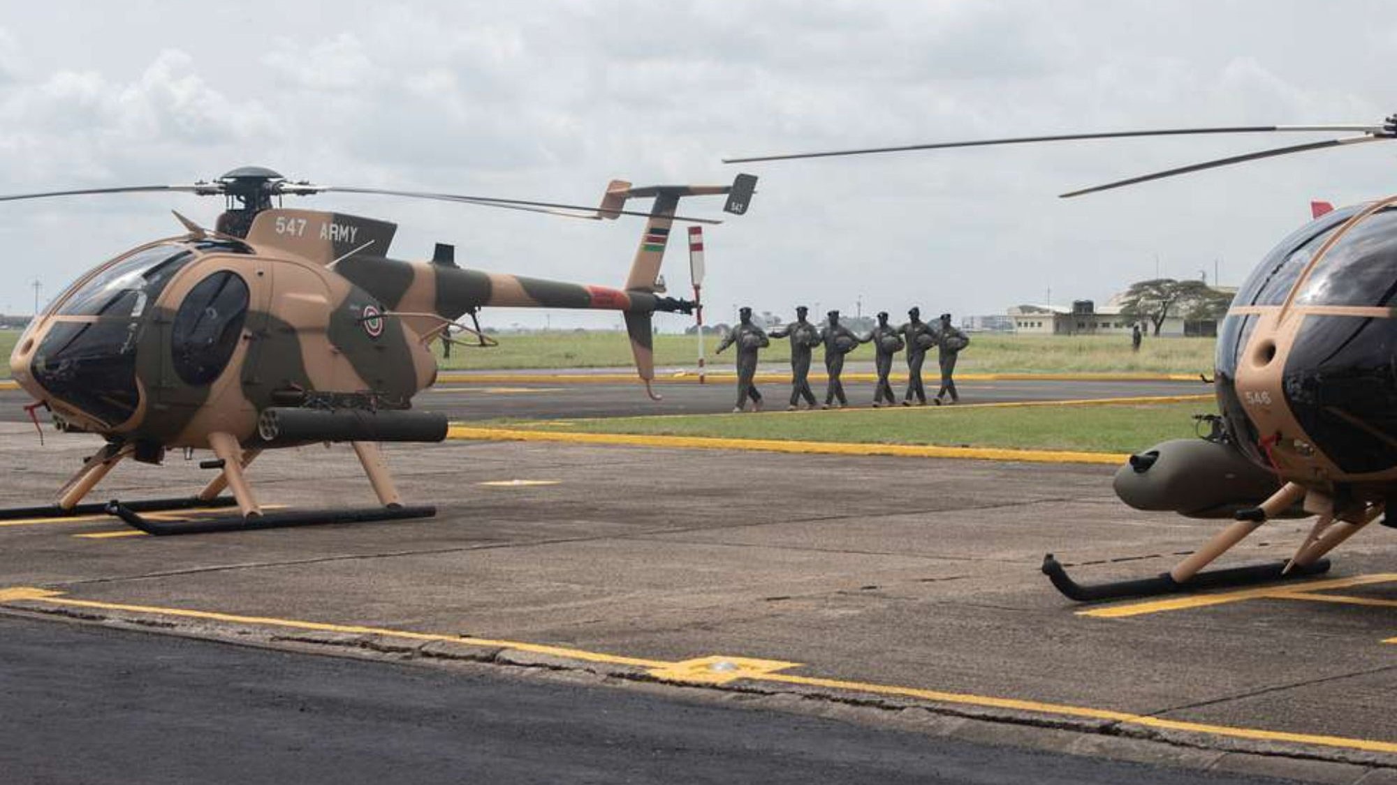 L'image montre une scène aérienne sur un terrain d'aviation. Deux hélicoptères, de couleur beige avec des motifs camouflage, sont stationnés sur le tarmac. À l'arrière-plan, un groupe de soldats en uniforme se dirige en formation vers un point de destination, créant une atmosphère dynamique et militaire. Le ciel est nuageux, ajoutant une ambiance dramatique à la scène. Le paysage autour est dégagé, avec des bâtiments épars visibles au loin.