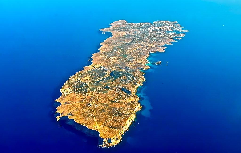 L'image représente une île vue du ciel, entourée par une vaste étendue d'eau bleue. L'île a une forme allongée et irrégulière, avec des côtes découpées et quelques zones rocheuses. Sa surface est en grande partie couverte de végétation, avec des teintes de jaune, de vert et de brun qui évoquent un terrain sec et accidenté. On peut également observer quelques petites baies et criques où l'eau semble se jeter sur le rivage. L'ensemble évoque une atmosphère de tranquillité et de beauté naturelle, typique d'une destination insulaire.