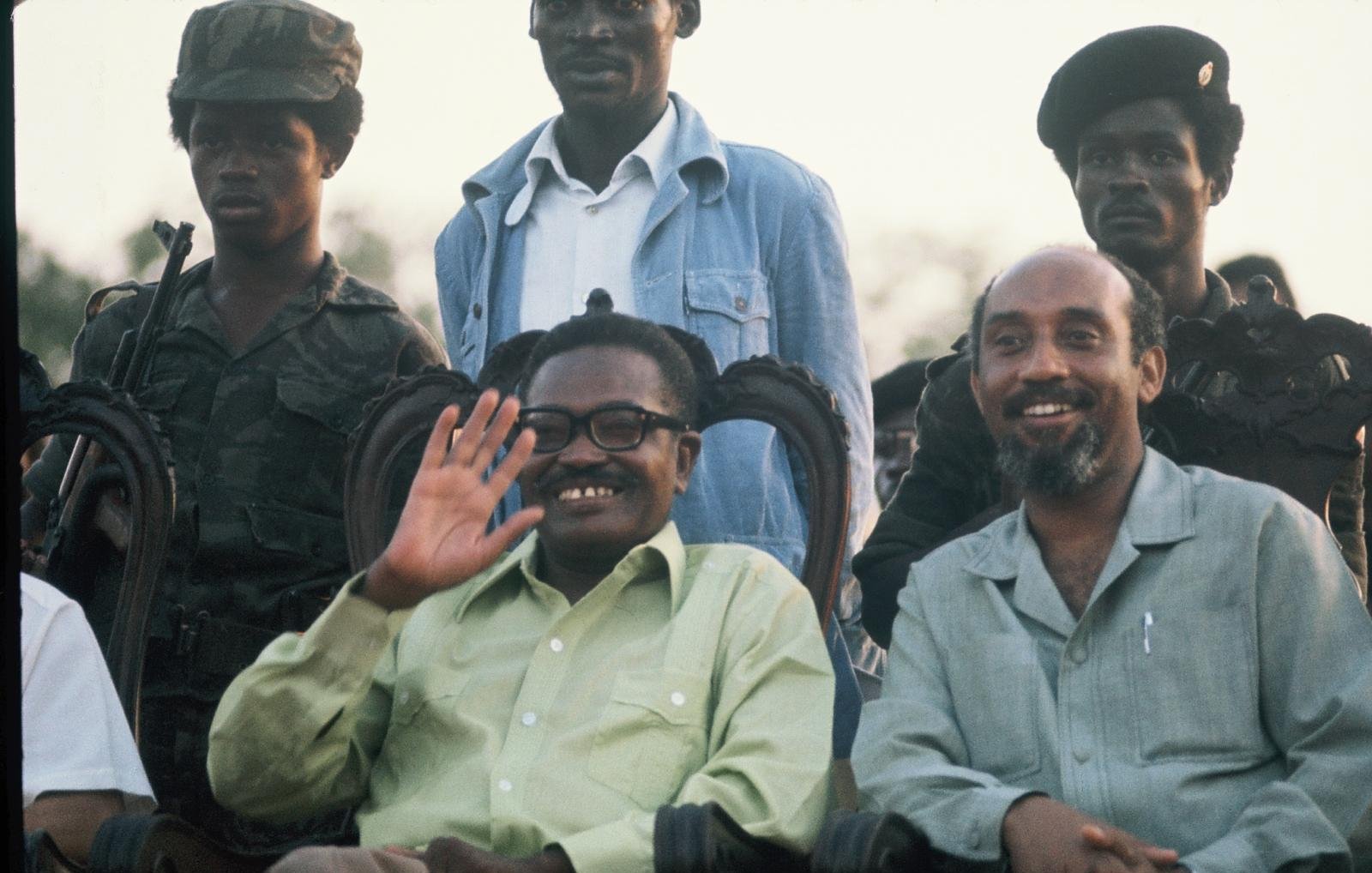 L'image présente un groupe de personnes, dont deux hommes au premier plan, assis sur des chaises ornées. L'un des hommes, souriant et portant des lunettes, salue de la main. Il a une chemise verte claire. À sa droite, un autre homme avec une chemise beige sourit également. En arrière-plan, plusieurs soldats en uniforme de camouflage et avec des casquettes se tiennent debout. L'environnement semble être en plein air, peut-être lors d'un événement officiel, avec un ciel illuminé par le crépuscule.