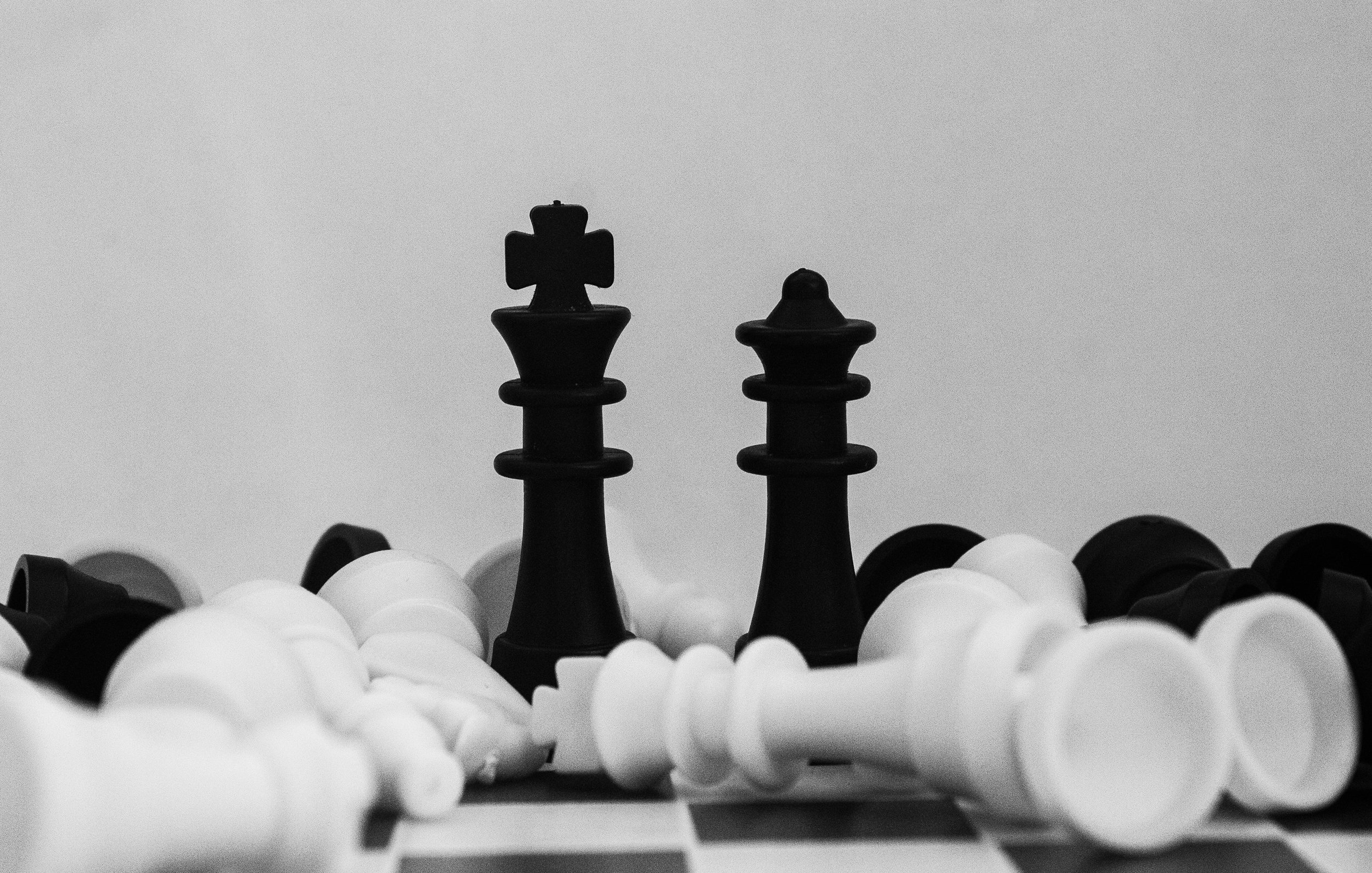 L'image présente deux pièces de jeu d'échecs, un roi noir et un roi blanc, se faisant face au centre. Ils sont placés sur un échiquier, entourés d'autres pièces éparpillées, à la fois noires et blanches. La composition est en noir et blanc, donnant une atmosphère sobre et artistique. Le fond est clair, contrastant avec les pièces sombres, ce qui met en valeur leur forme et détails. L'ensemble évoque une scène de stratégie et de confrontation dans le jeu d'échecs.