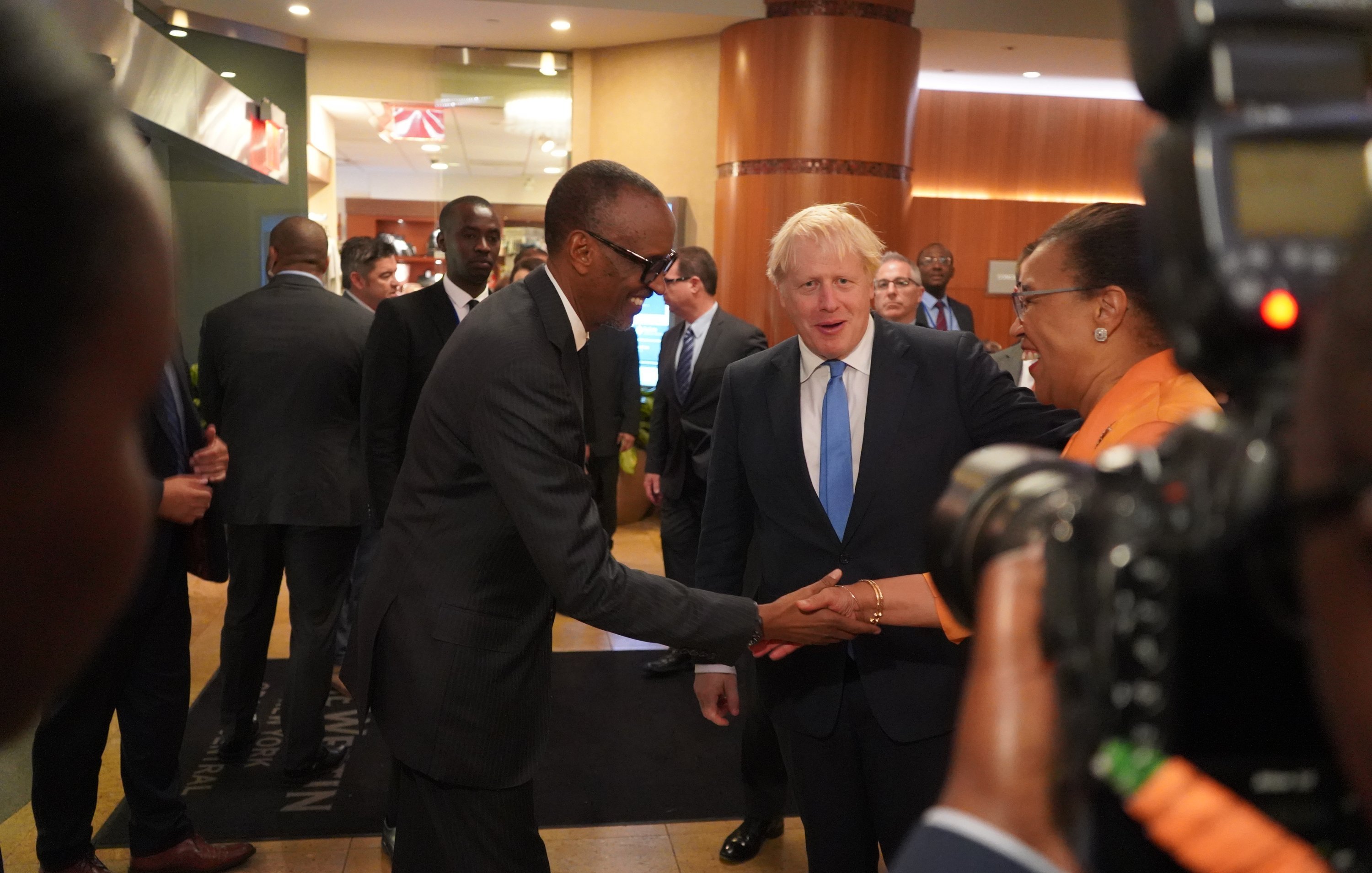 L'image représente une scène dans un hall d'hôtel ou de conférence, où plusieurs personnes sont rassemblées. Au centre, deux hommes se serrent la main : l'un est de grande taille, portant un costume sombre et des lunettes, tandis que l'autre, avec des cheveux blonds, est habillé d'un costume clair. Ils affichent tous deux des sourires amicaux. En arrière-plan, on peut voir plusieurs autres personnes en discussion, ainsi que des détails du décor intérieur, comme des murs aux tons chauds et des éléments de lumière. L'ambiance semble professionnelle et conviviale.