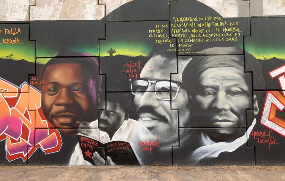 Cette image montre une fresque murale vibrante et colorée qui rend hommage à des figures importantes de l'histoire africaine. Au centre, il y a des portraits de trois personnes, chacun portant une expression sérieuse et réfléchie. Les visages sont peints en noir et blanc, mettant en valeur les détails de leurs traits. À gauche, un homme sourit légèrement, symbolisant l'espoir et la détermination. Au centre, un homme barbu porte des lunettes et affiche un regard intense, représentant la sagesse. À droite, un homme au visage marqué par le temps semble contemplatif, illustrant la profondeur de la pensée. En arrière-plan, des motifs géométriques et des couleurs riches ajoutent une dynamique à l'ensemble. Des inscriptions en lettres blanches ponctuent la fresque, partageant des pensées inspirantes sur l'identité, la culture et l'impact de ces figures dans le monde. Cette œuvre évoque une ambiance de fierté et d'émancipation culturelle.