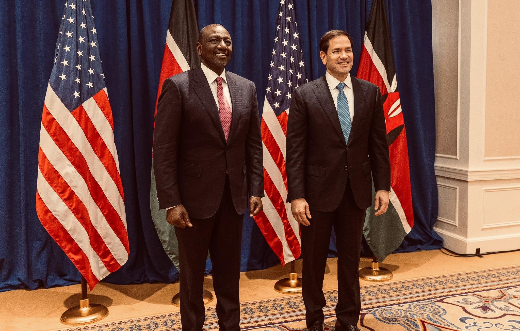 Sur cette image, nous voyons deux hommes debout côte à côte, souriants. À gauche, un homme portant un costume noir et une cravate rouge. Il a la peau foncée et une posture confiante. À droite, un homme avec un costume sombre et une cravate bleu clair. Il a la peau plutôt claire et un sourire engageant. Derrière eux, il y a trois drapeaux : deux des États-Unis, avec des étoiles blanches sur fond bleu et stripes rouges et blancs, et un drapeau du Kenya, qui a des bandes horizontales noires, rouges et vertes, avec un bouclier au centre. Le décor est élégant, avec un fond bleu et un tapis orné à leurs pieds. L'atmosphère semble formelle et amicale.