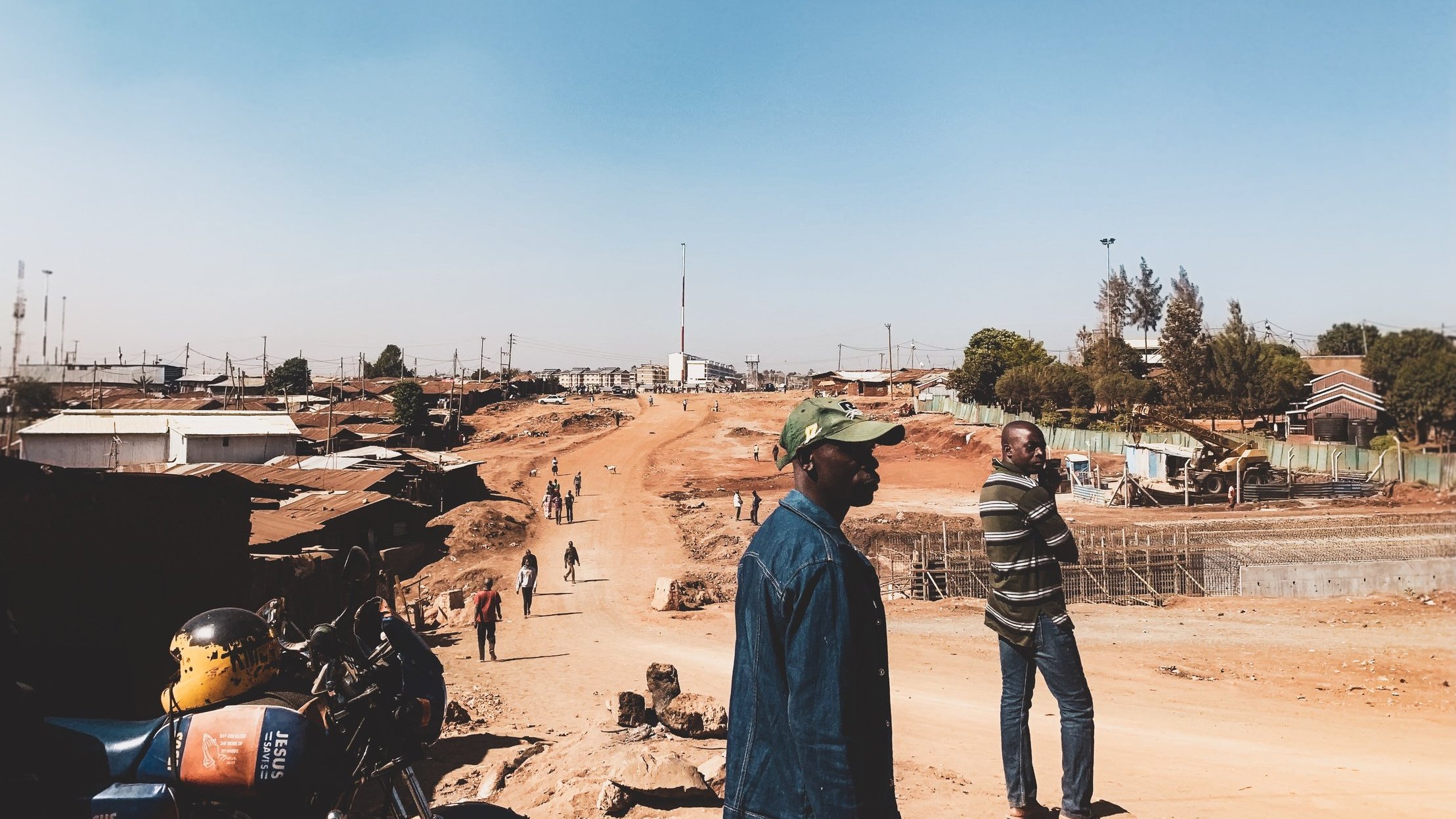 Dans cette image, nous voyons une scène d'une rue en construction dans une zone urbaine. Le ciel est dégagé et d'un bleu clair. Au premier plan, deux hommes se tiennent debout : l'un porte une veste en jean et une casquette, tandis que l'autre est vêtu d'un sweater rayé. À droite, on aperçoit un chemin de terre, entouré de bâtiments en dur et d'habitations simples. À gauche, une moto est garée, et plusieurs personnes se déplacent le long de la route. L'atmosphère semble calme, mais l'environnement indique une activité en cours avec des travaux de construction en arrière-plan.