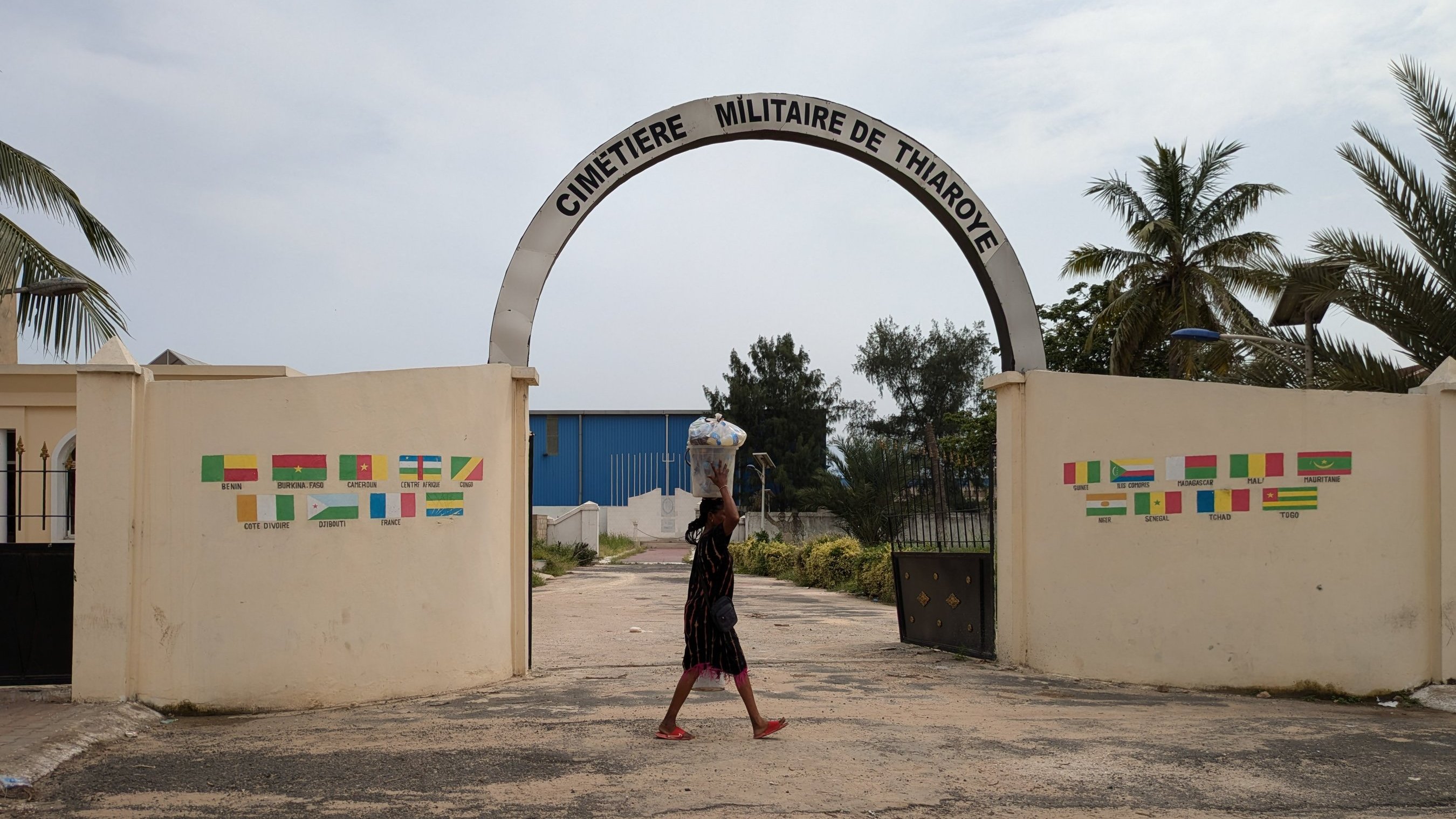 L'image montre l'entrée du cimetière militaire de Thiaroye. Un grand arc de portail est inscrit avec le nom "CIMETIÈRE MILITAIRE DE THIAROYE". En arrière-plan, on peut apercevoir quelques arbres, dont des palmiers, qui ajoutent une touche tropicale à la scène. Au premier plan, une personne marche, portant un grand récipient sur sa tête. Le sol est en béton, avec des traces de circulation. De part et d'autre de l'entrée, des murs affichent des drapeaux de différentes nations, apportant une sensation de diversité. L'ensemble dégage une ambiance calme et respectueuse.